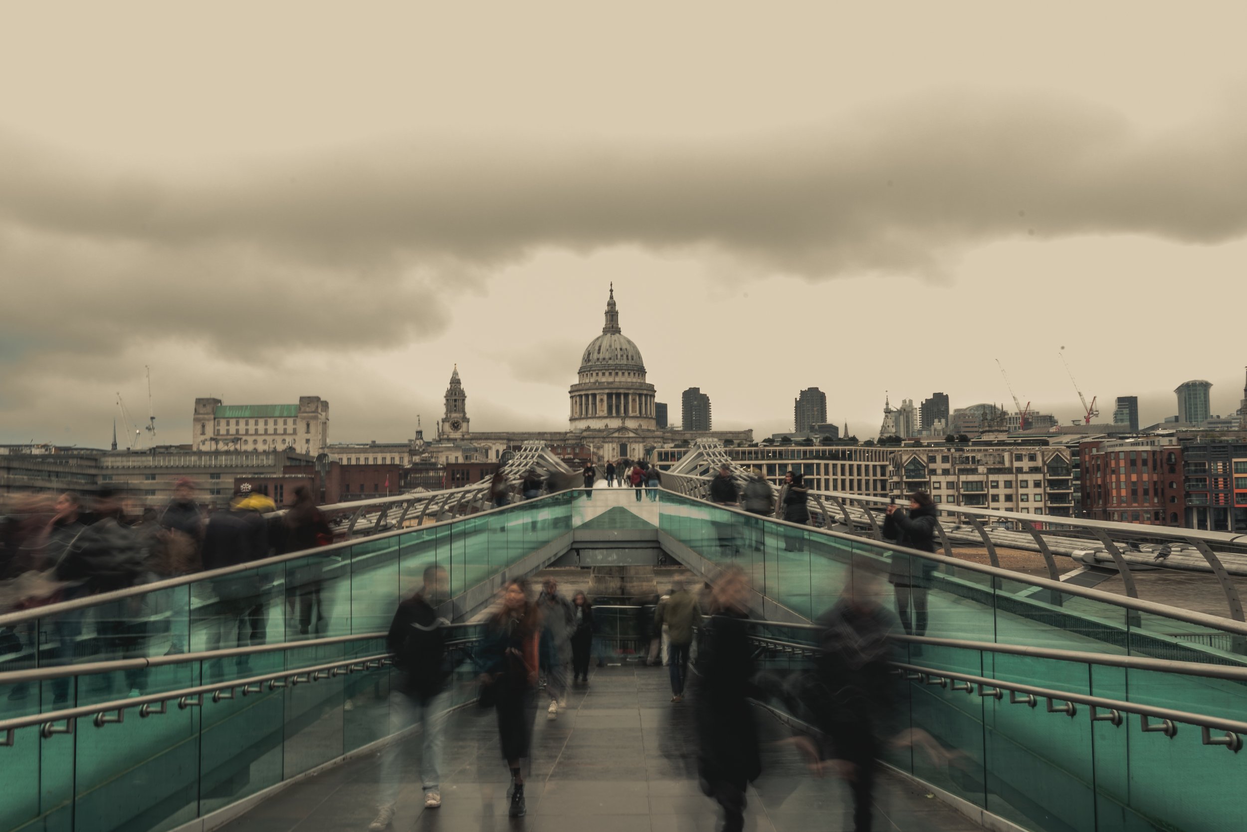 View of Millennium Bridge in London with St. Paul's Cathedral in the background, cloudy sky, and blurred pedestrians walking on the bridge.