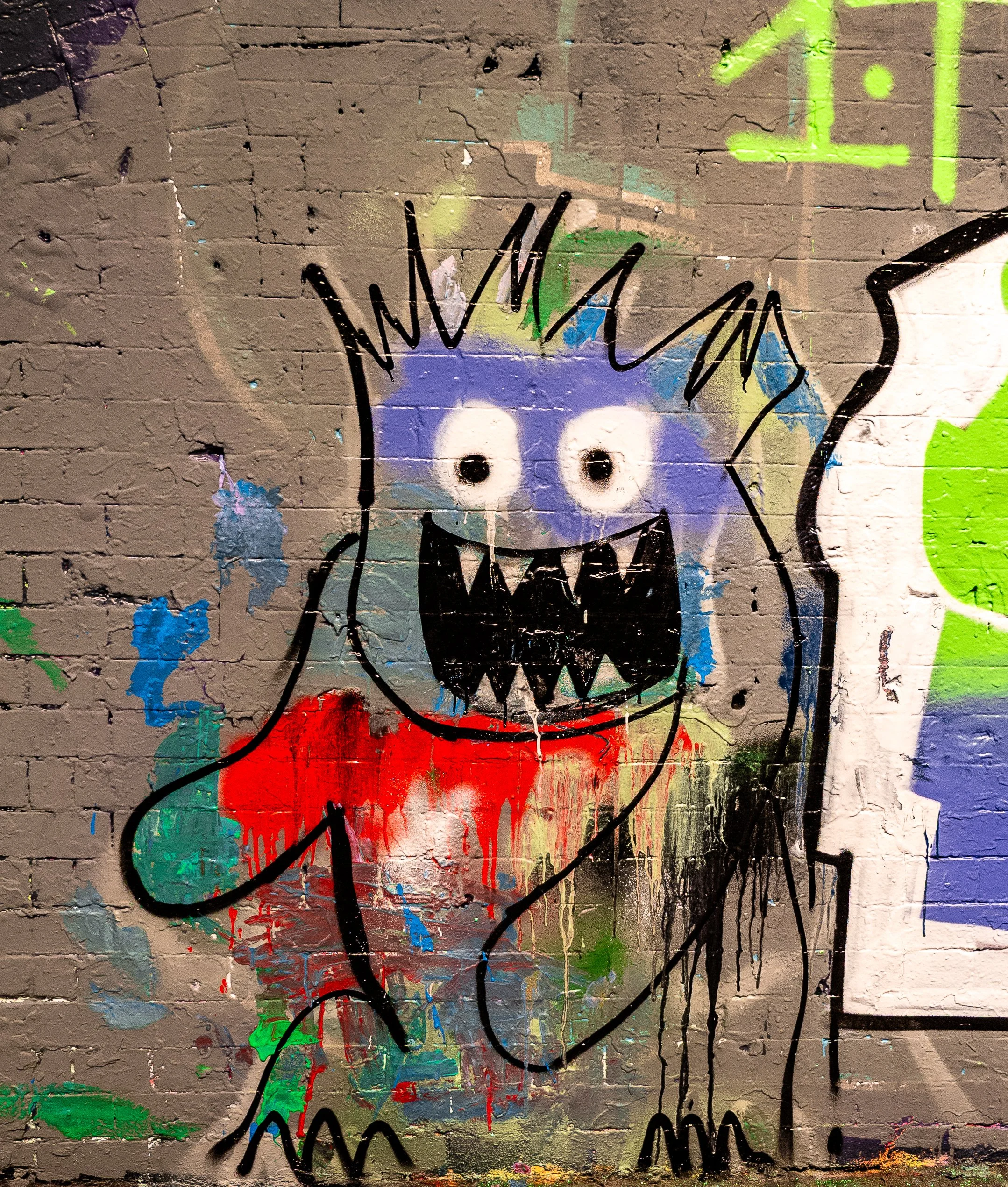 Street art graffiti of a cartoon monster with a round face, big eyes, sharp teeth, and spiky hair, painted on a brick wall with colorful drips and splashes.