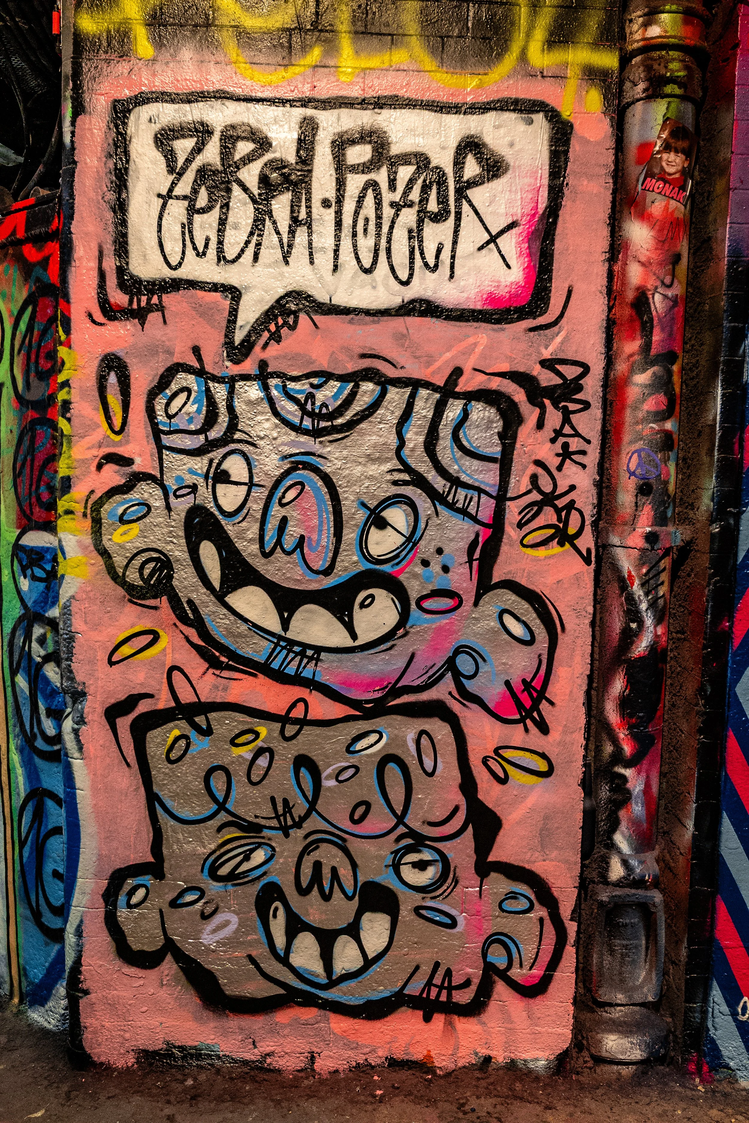 Colorful street art graffiti featuring two cartoon-like monster faces with exaggerated features, surrounded by tags and symbols in various colors.