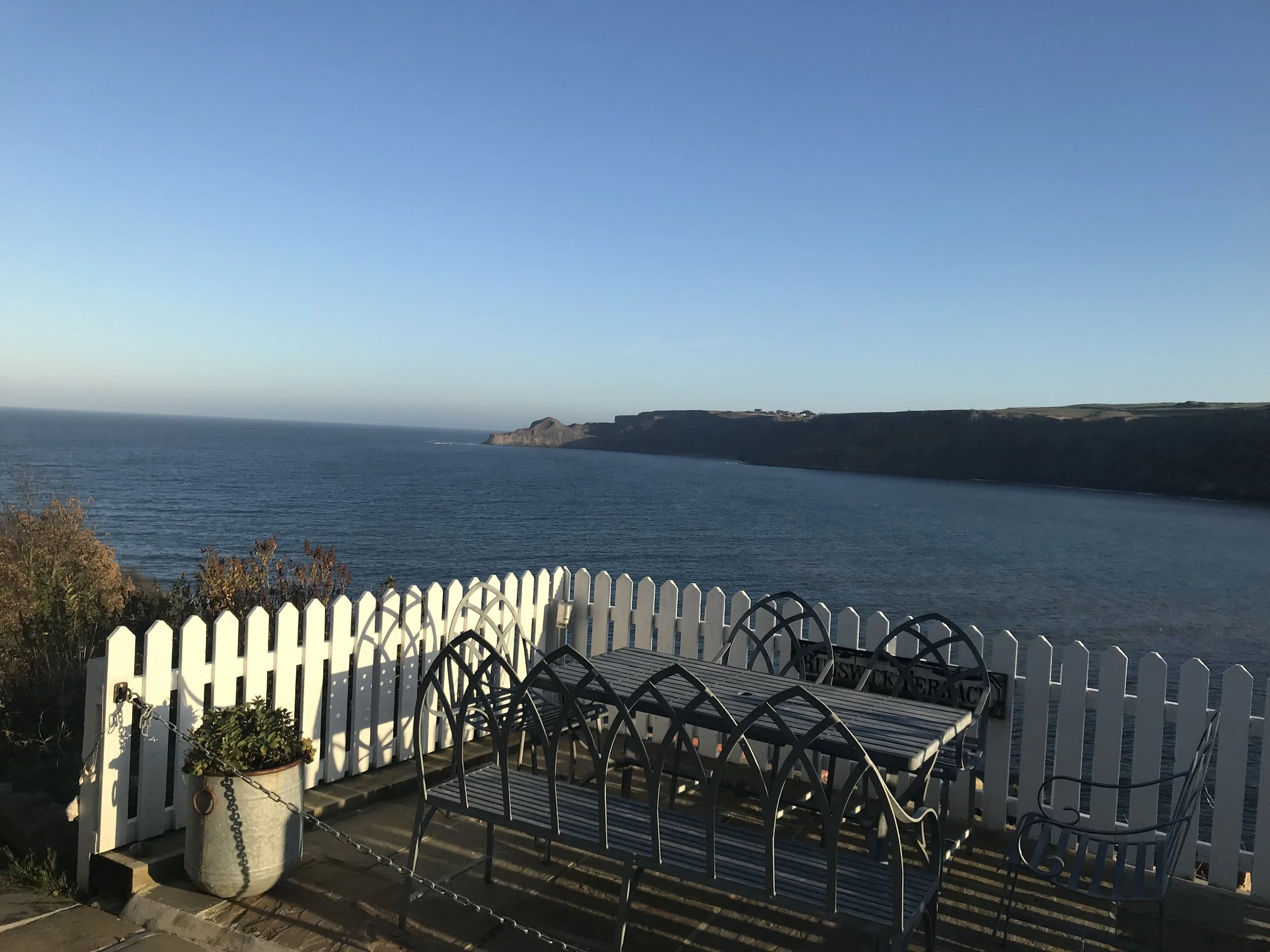 A seaside deck with white picket fence and black metal outdoor furniture, overlooking calm ocean waters with cliffs in the distance under a clear blue sky.