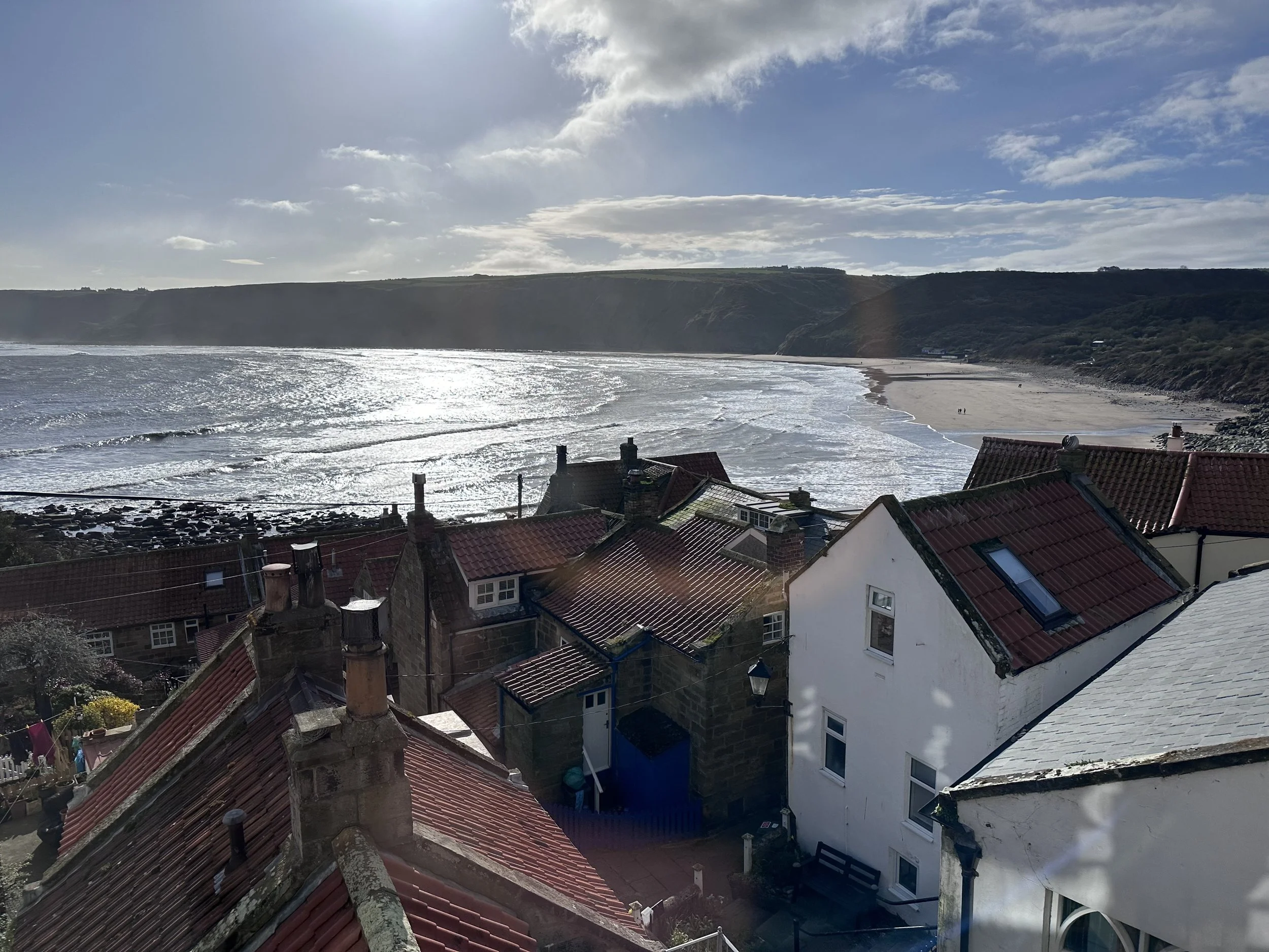 A view of a bay or ocean with waves reflecting the sunlight, a sandy beach, and houses with red and gray roofs overlooking the shore, with a hill covered in greenery in the background.