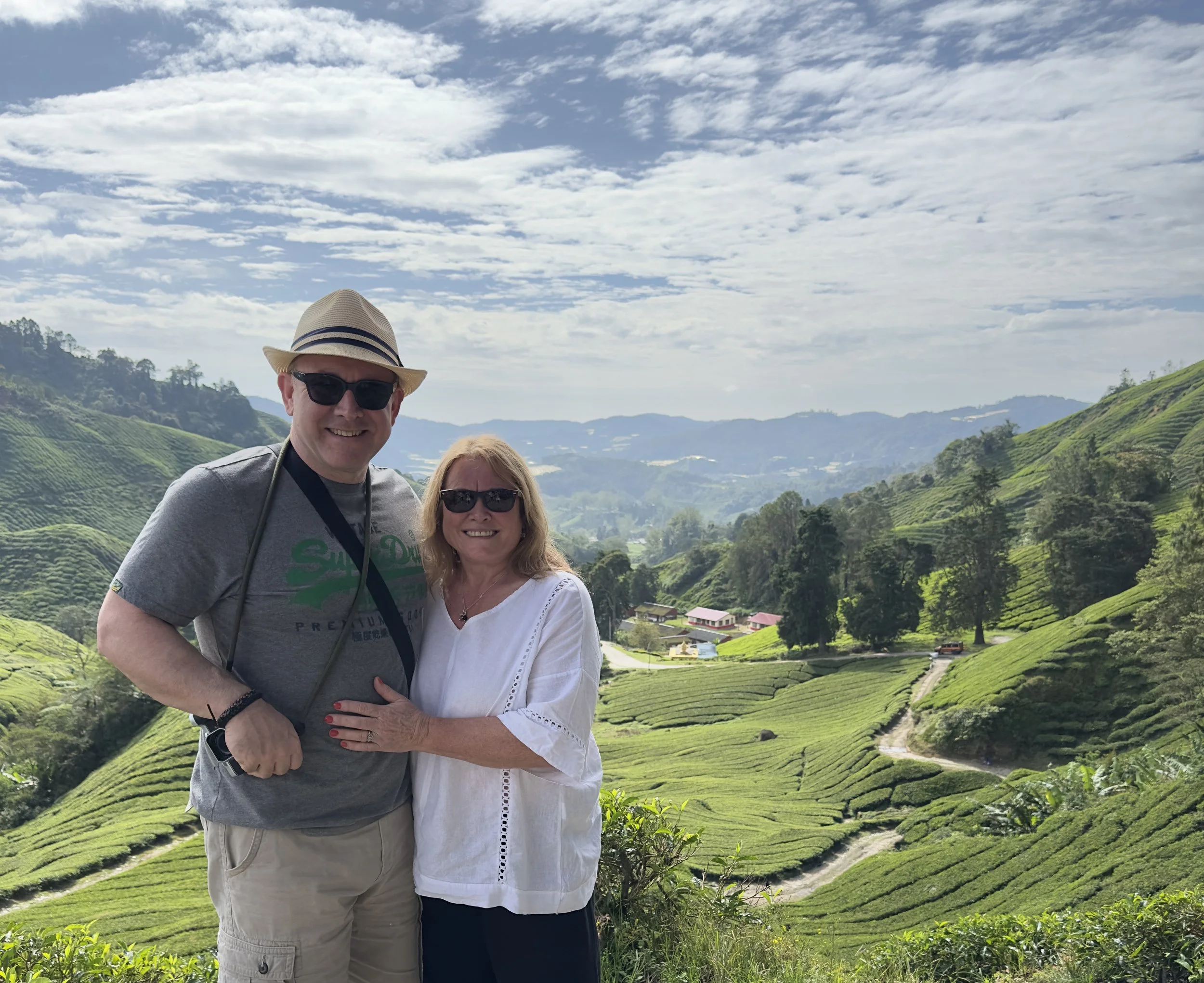 A smiling couple stands close together, posing for a photo in a lush green hilly landscape under a partly cloudy sky. The man is wearing a fedora hat, sunglasses, a gray t-shirt, and khaki shorts with a camera strap around his neck. The woman is wear