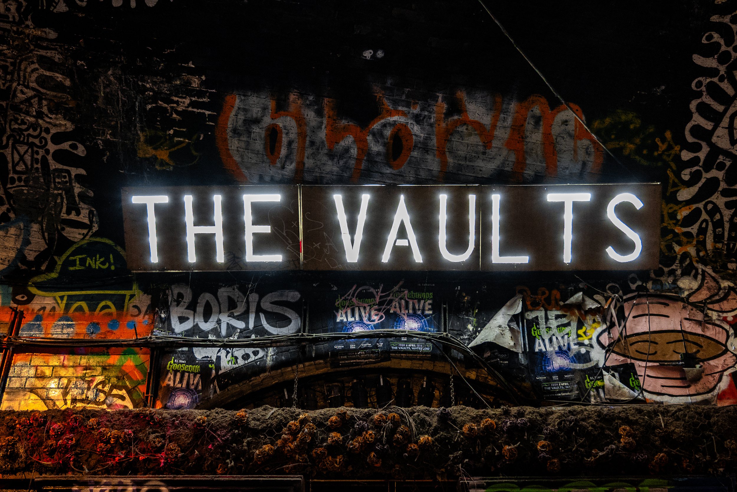 Neon sign reading 'THE VAULTS' in front of a graffitied wall.