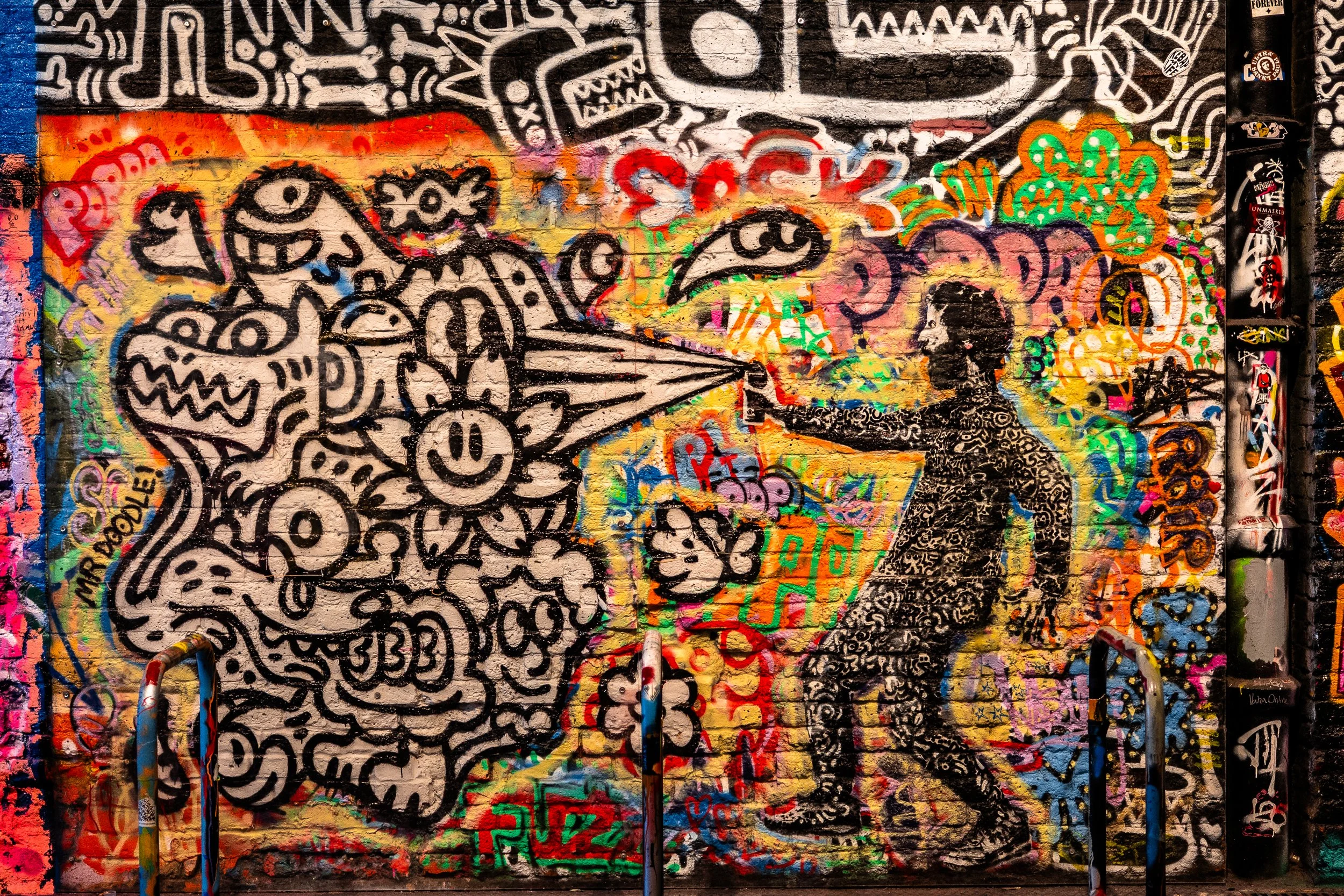 Colorful graffiti mural on a brick wall depicting a person with curly hair shooting a spray of paint at a vibrant, abstract, cartoon-style creature with a large head, sharp teeth, and various expressive faces, surrounded by various graffiti tags and 