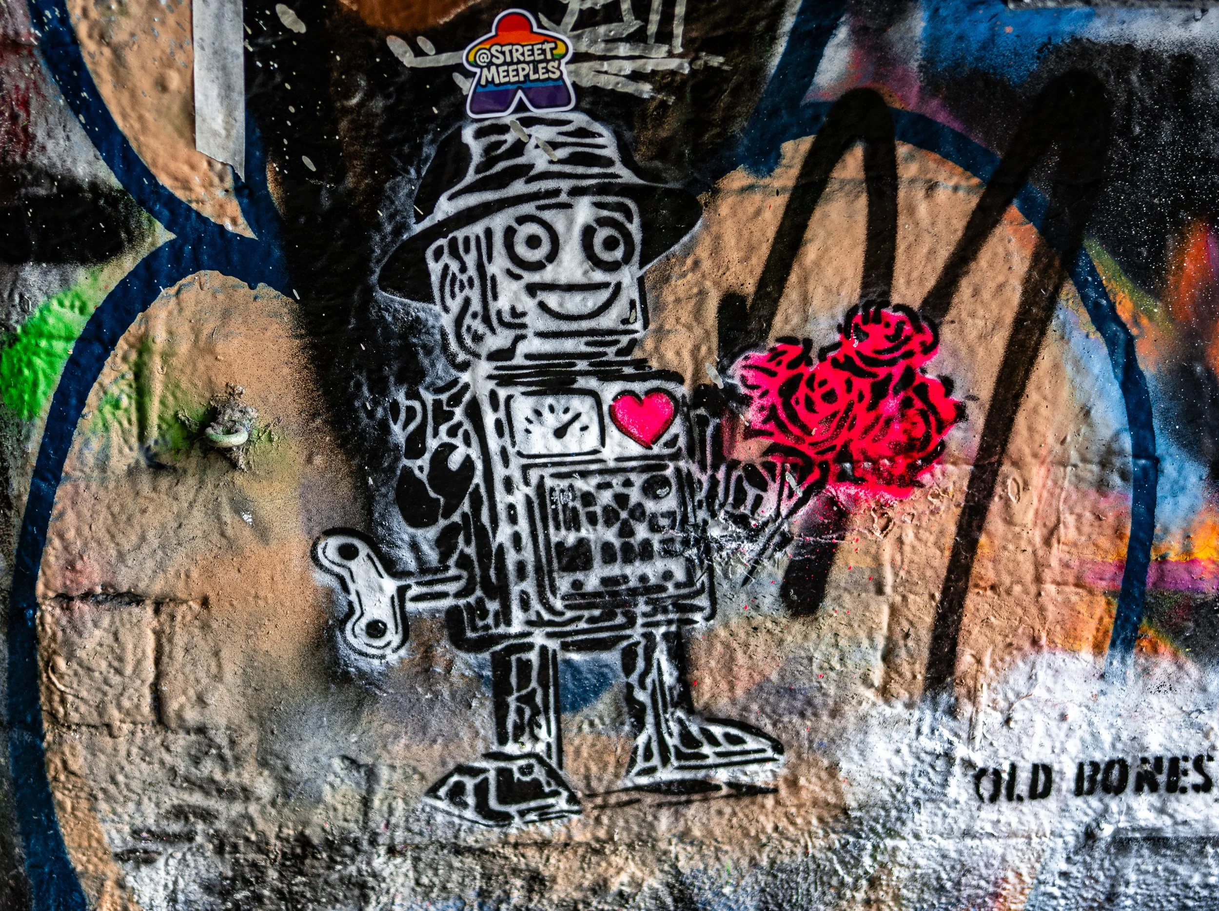 Street art graffiti of a robot figure with a smiling face, wearing a wizard hat, holding a bunch of red roses in one hand and a small heart on its chest, on a textured wall with various colors and tags. The text 'OLD BONES' appears in the bottom righ