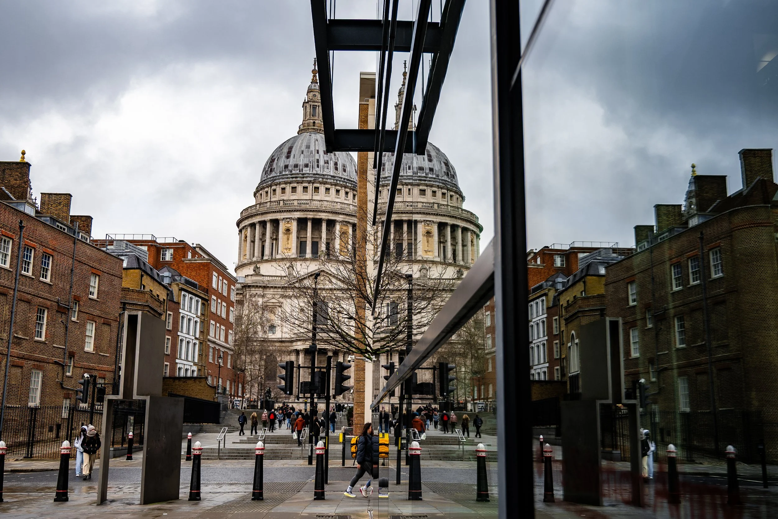 Reflection of St. Paul's Cathedral in a glass building on a city street with pedestrians, during cloudy weather.