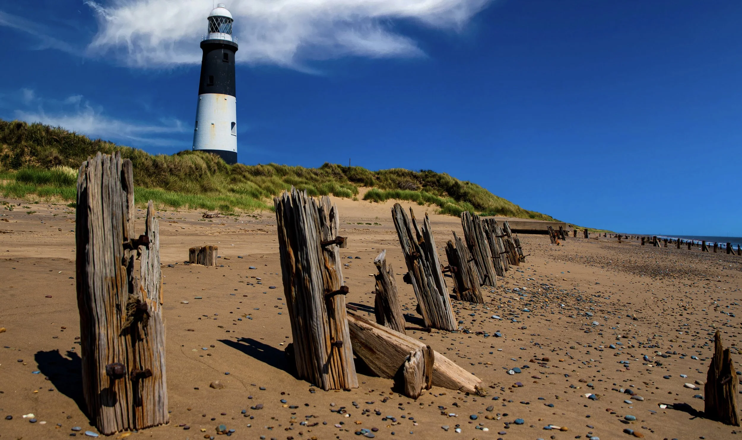 Beach with old wooden posts and a lighthouse in the background under a blue sky.