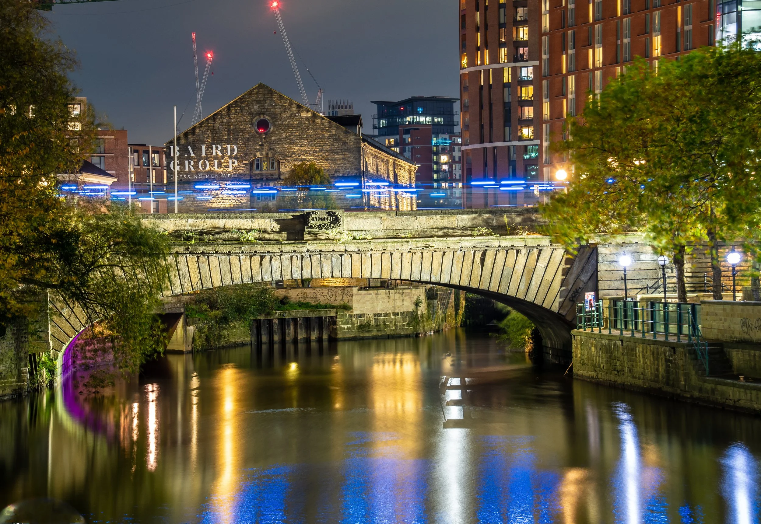 Nighttime cityscape featuring a stone bridge over a river, illuminated buildings, and construction cranes in the background.
