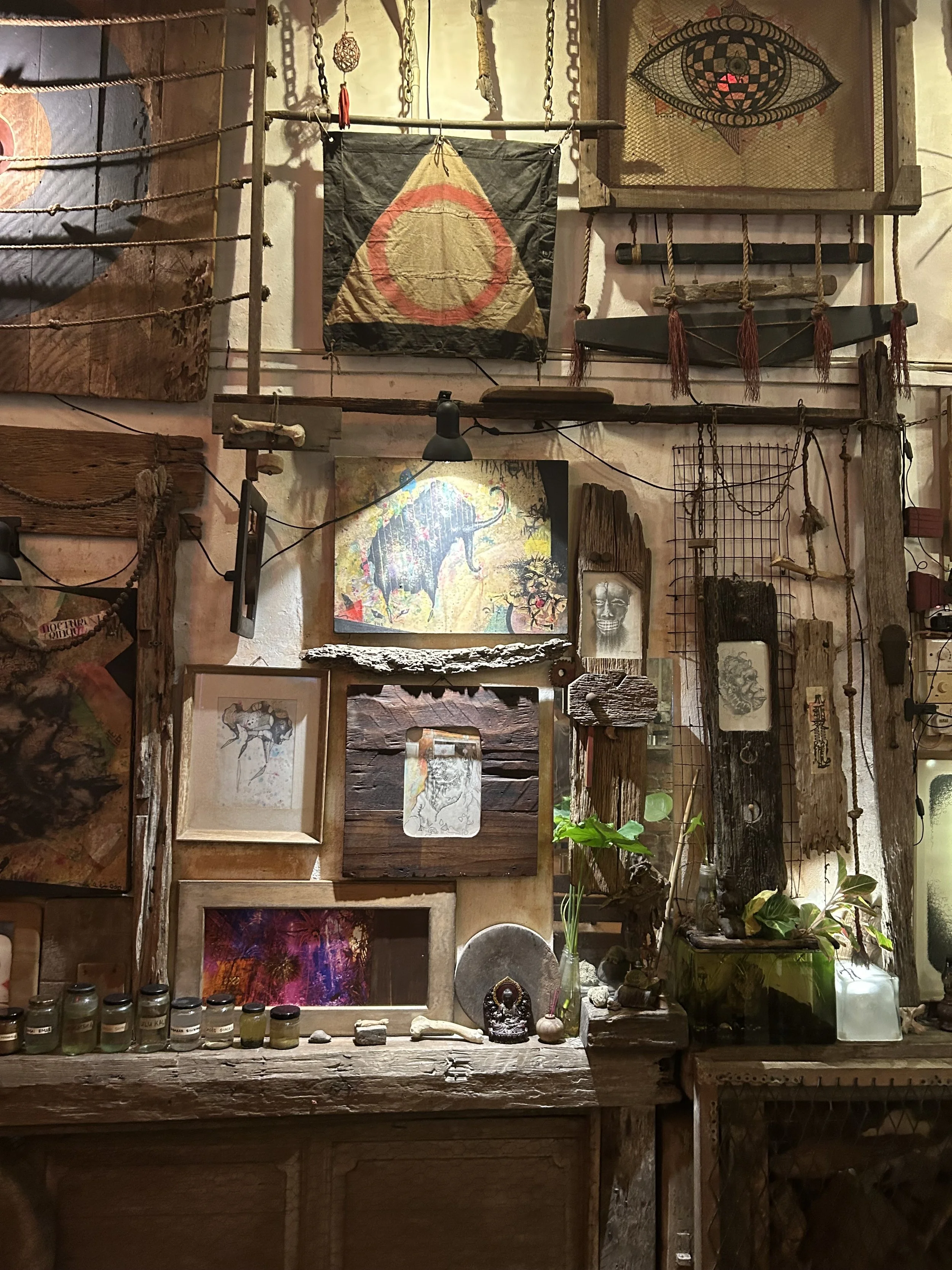 Eclectic wall decorated with framed artwork, wooden and metal objects, plants, and artwork including a flag with a circle and triangle design. Various art pieces, plants, and decorative items are arranged on a wooden surface and hanging on the wall.