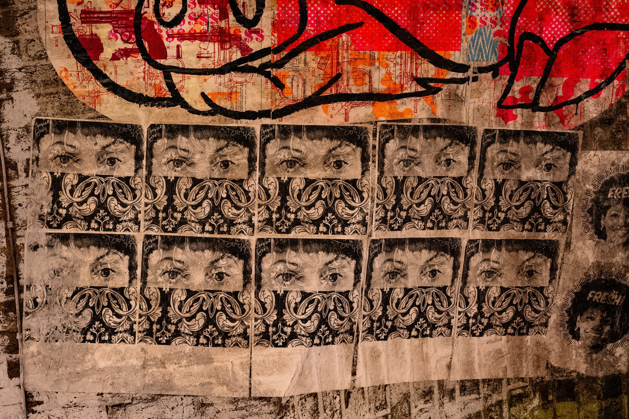 Black and white posters of a person's eyes framed by ornate designs, arranged in a grid on a concrete wall with spray-painted and sticker graffiti.