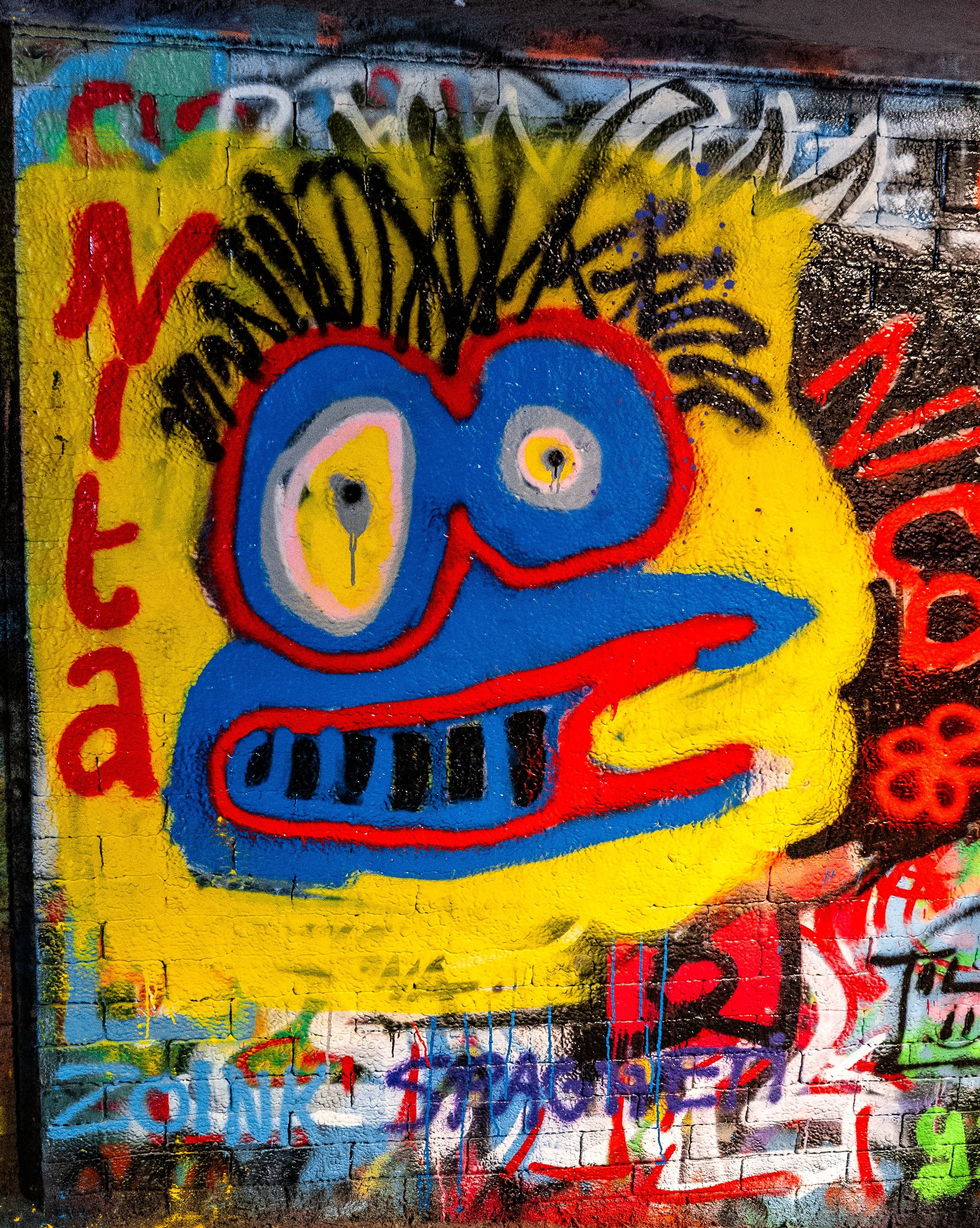 Graffiti art of a colorful cartoon-like face with yellow background, large blue eyes outlined in red, black eyebrows, and a blue mouth with red lips on a brick wall.