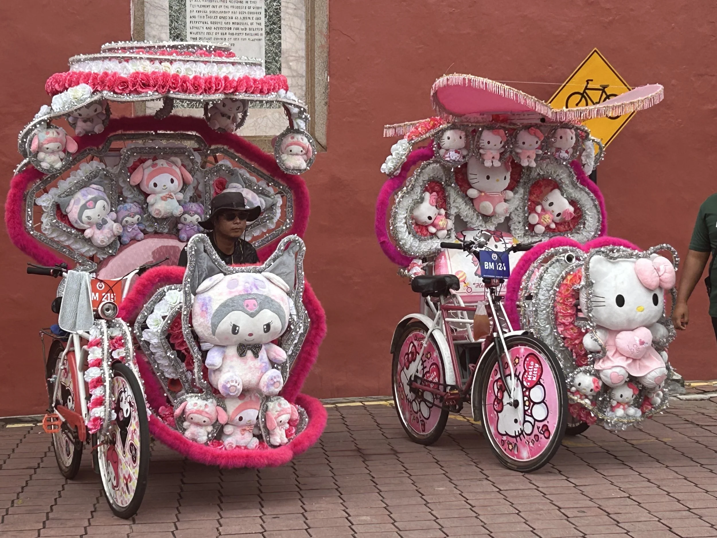 Two decorated bicycles featuring Hello Kitty and other plush toys, with pink, silver, and white embellishments, parked on a brick sidewalk against a reddish wall.