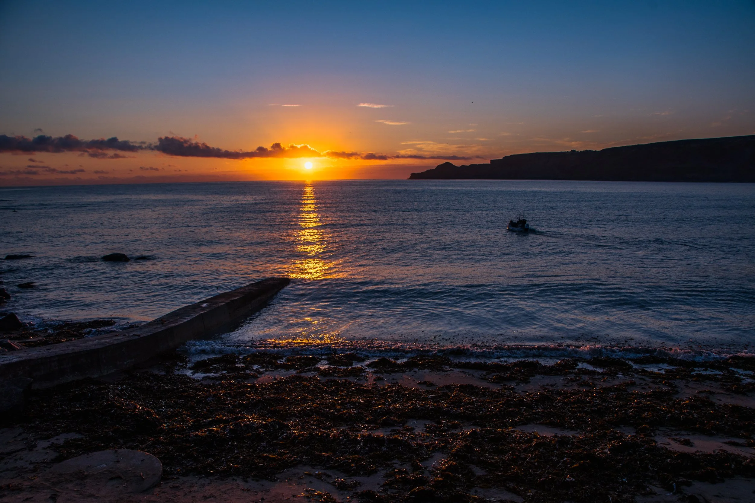 Sunset over the ocean with a small boat and rocky shoreline in the foreground.