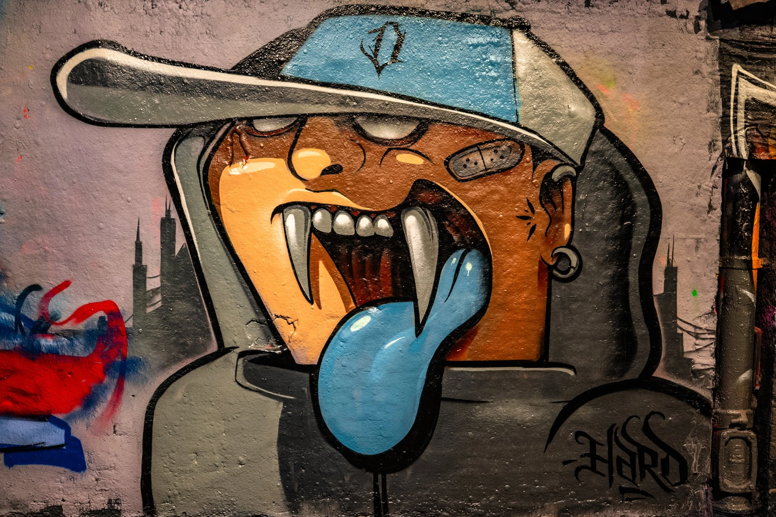 Colorful graffiti art of a creature with a large open mouth showing sharp teeth and a long blue tongue, wearing a baseball cap and an earring, on a concrete wall.