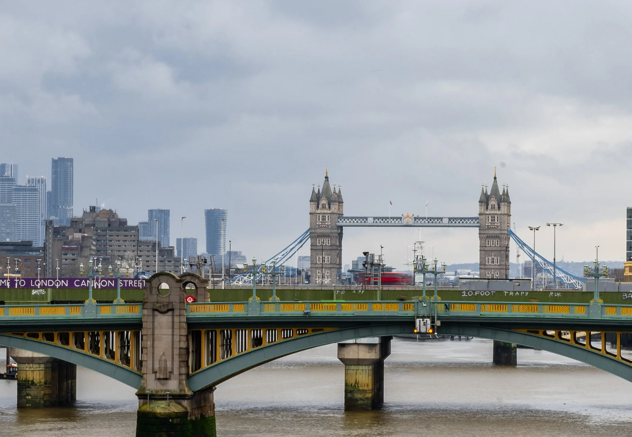 View of Tower Bridge in London with a bridge in the foreground spanning the River Thames, and the city skyline in the background under cloudy skies.