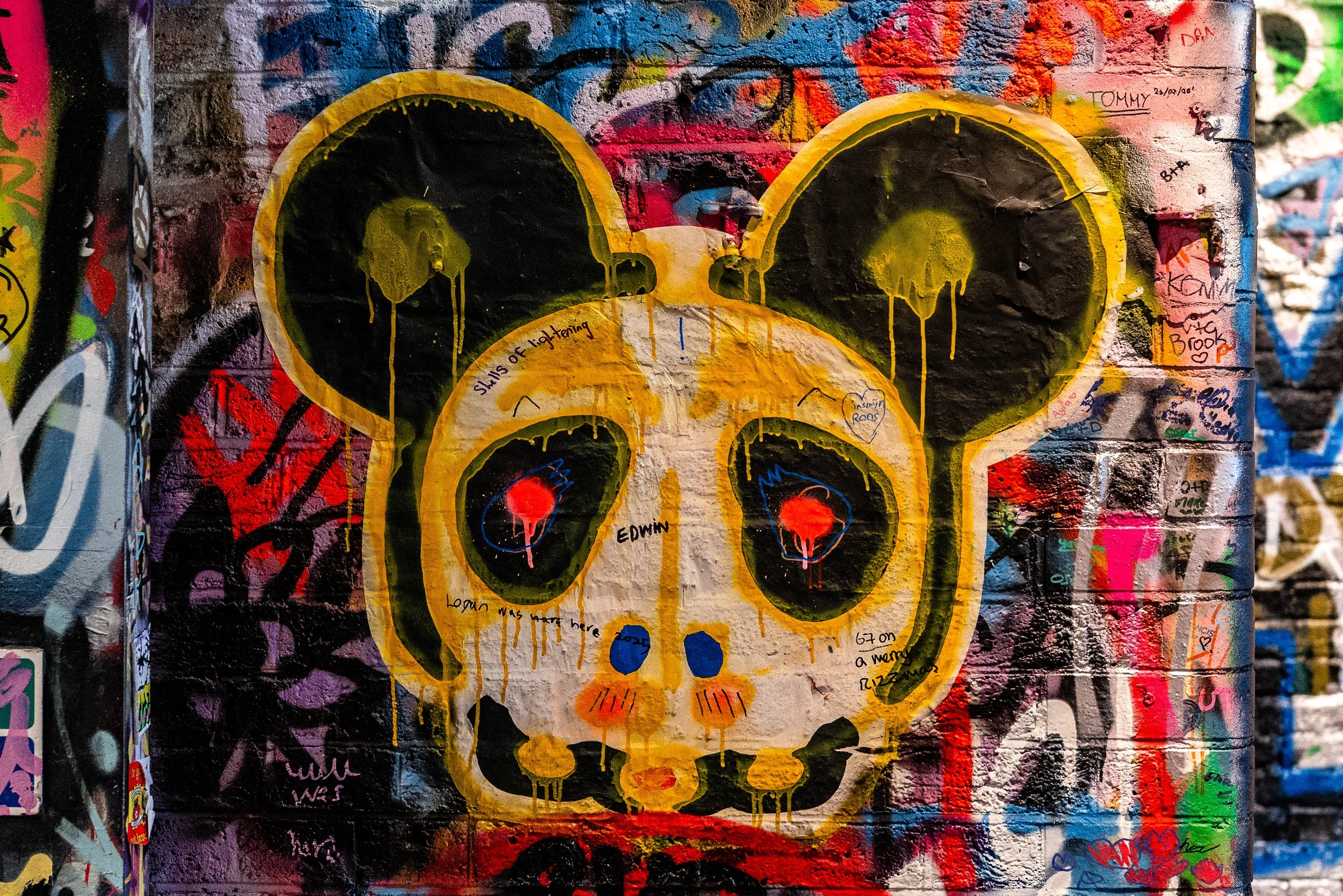 Colorful graffiti mural of a stylized mouse skull with yellow, black, and orange colors, dripping paint, and handwritten notes on a brick wall.