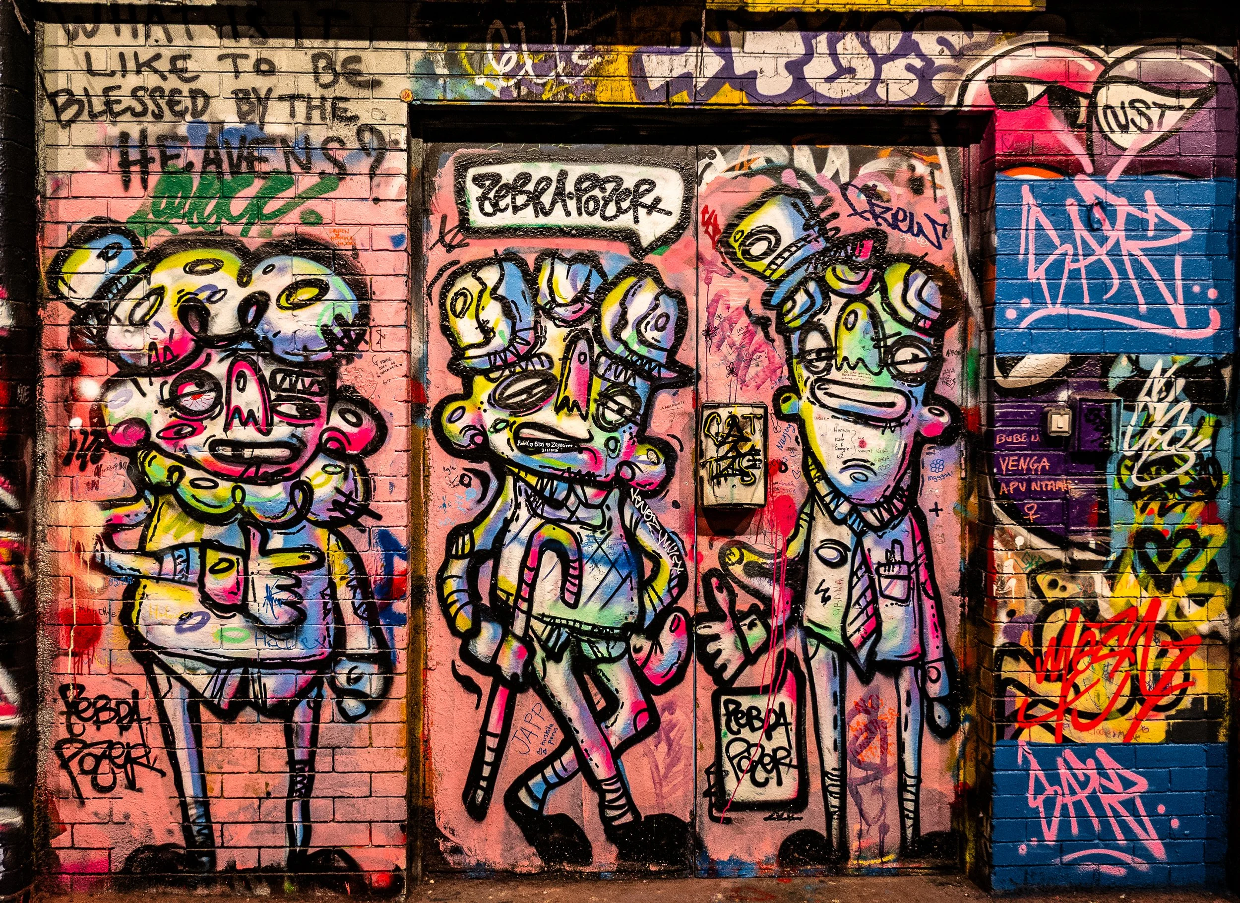 Colorful street art mural on brick wall showing cartoonish characters with exaggerated features, graffiti tags, and various vibrant colors including pink, blue, yellow, and black.
