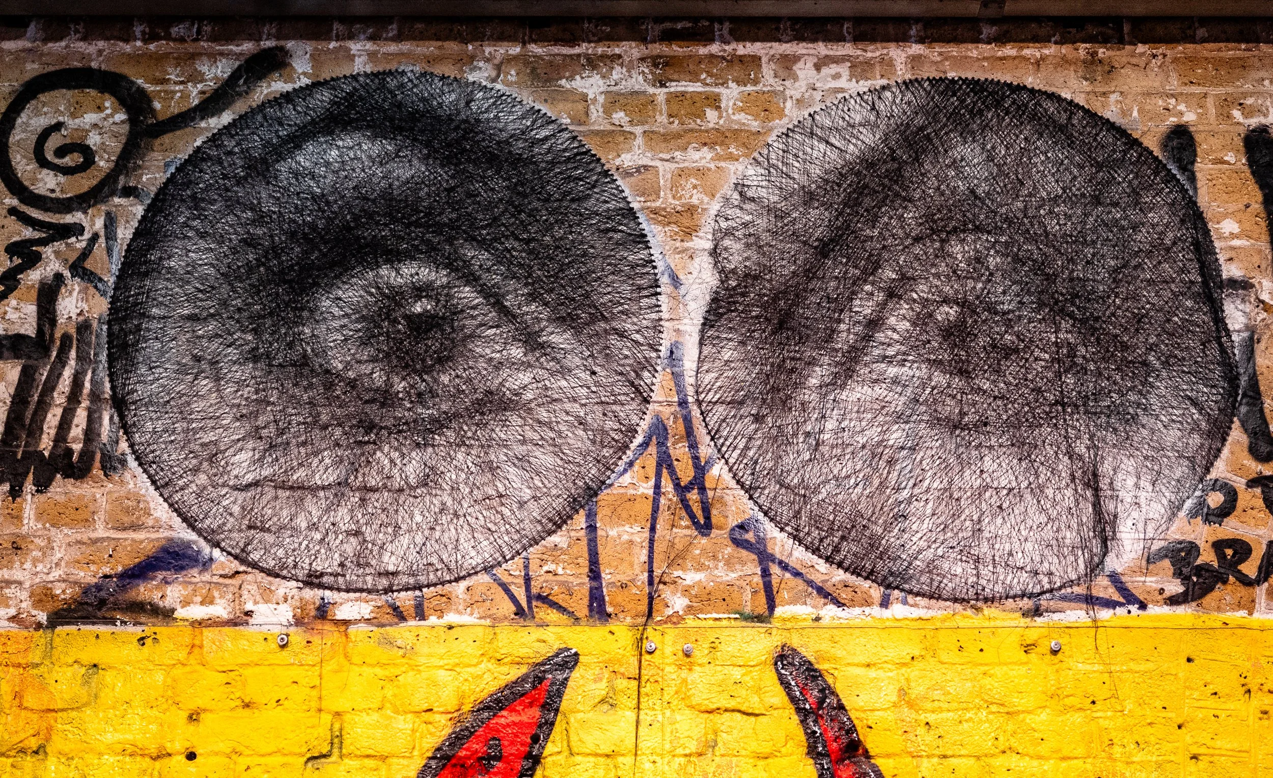 Street art featuring two black circular wire mesh sculptures on a brick wall, with colorful graffiti and yellow paint at the bottom.