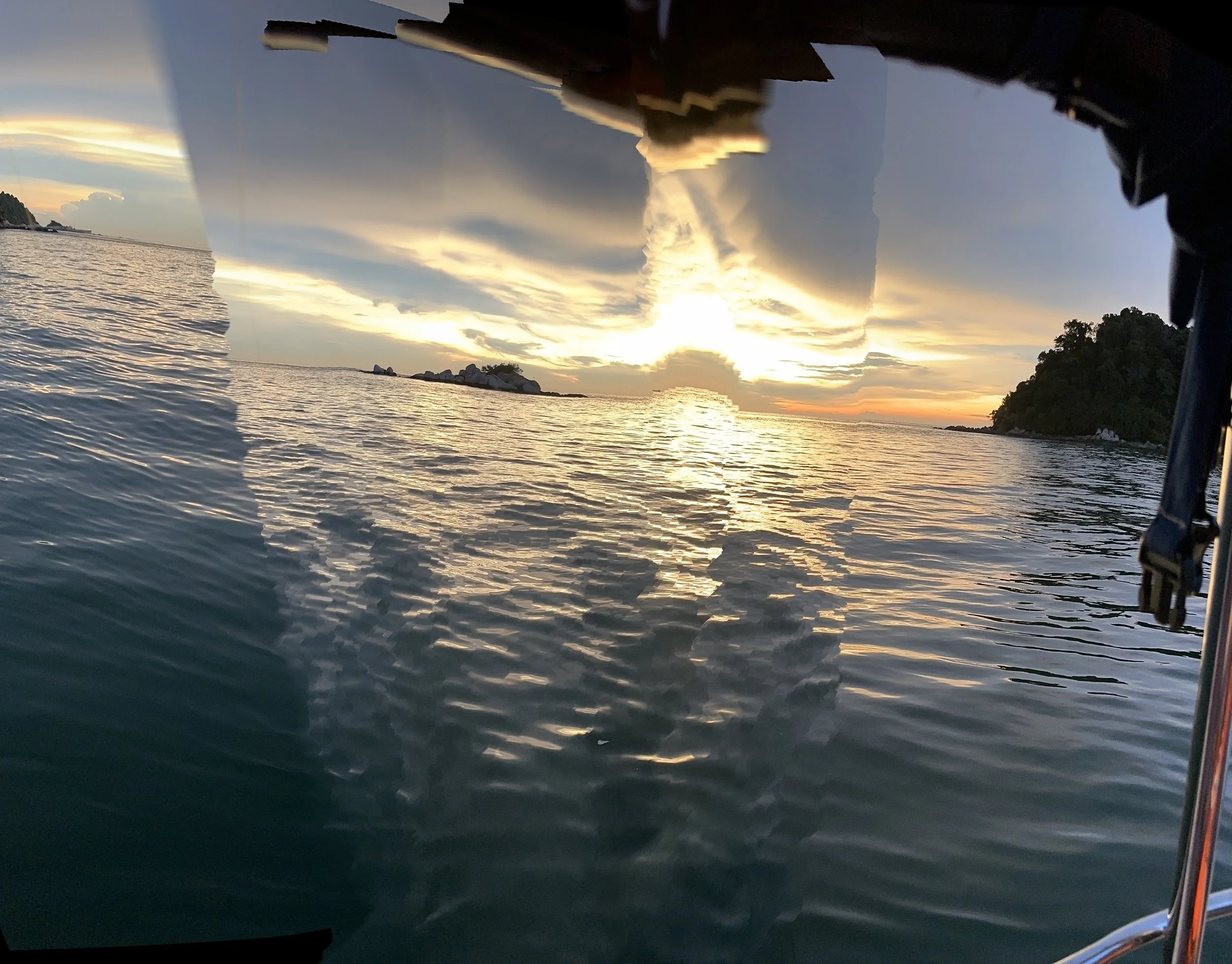 Sunset over the ocean with reflections on the water, view from under a boat's hull showing parts of the boat and a distant island.