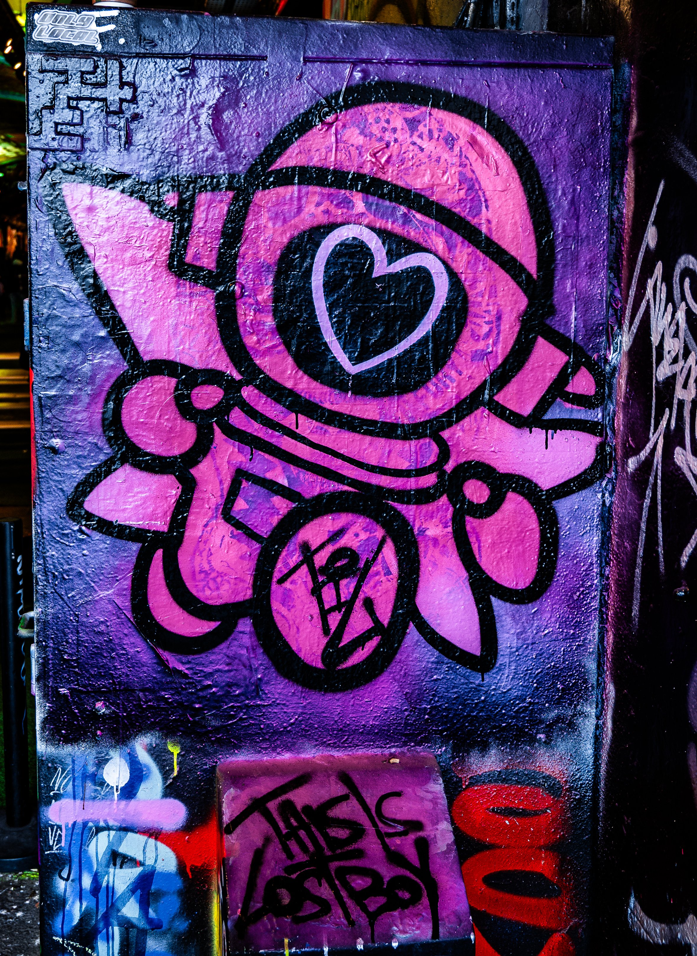 Pink and purple graffiti of an astronaut with a heart on the helmet, holding a flag and a shield, with various tags and graffiti on a wall.