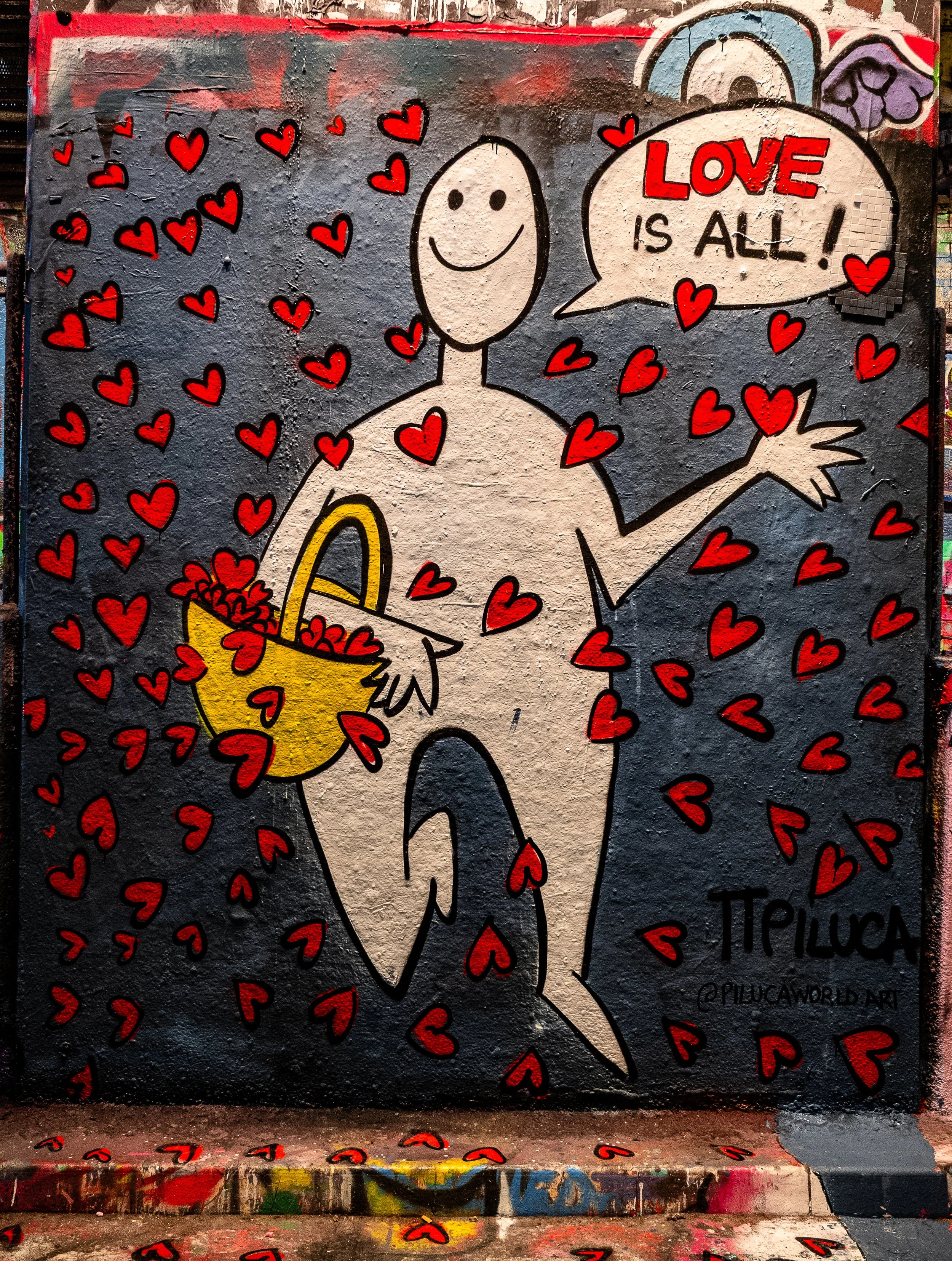 Street art mural of a smiling, nude, cartoon-style figure holding a yellow basket overflowing with red hearts. The figure has a speech bubble that says, 'LOVE IS ALL!'. The background is dark with red hearts scattered throughout.