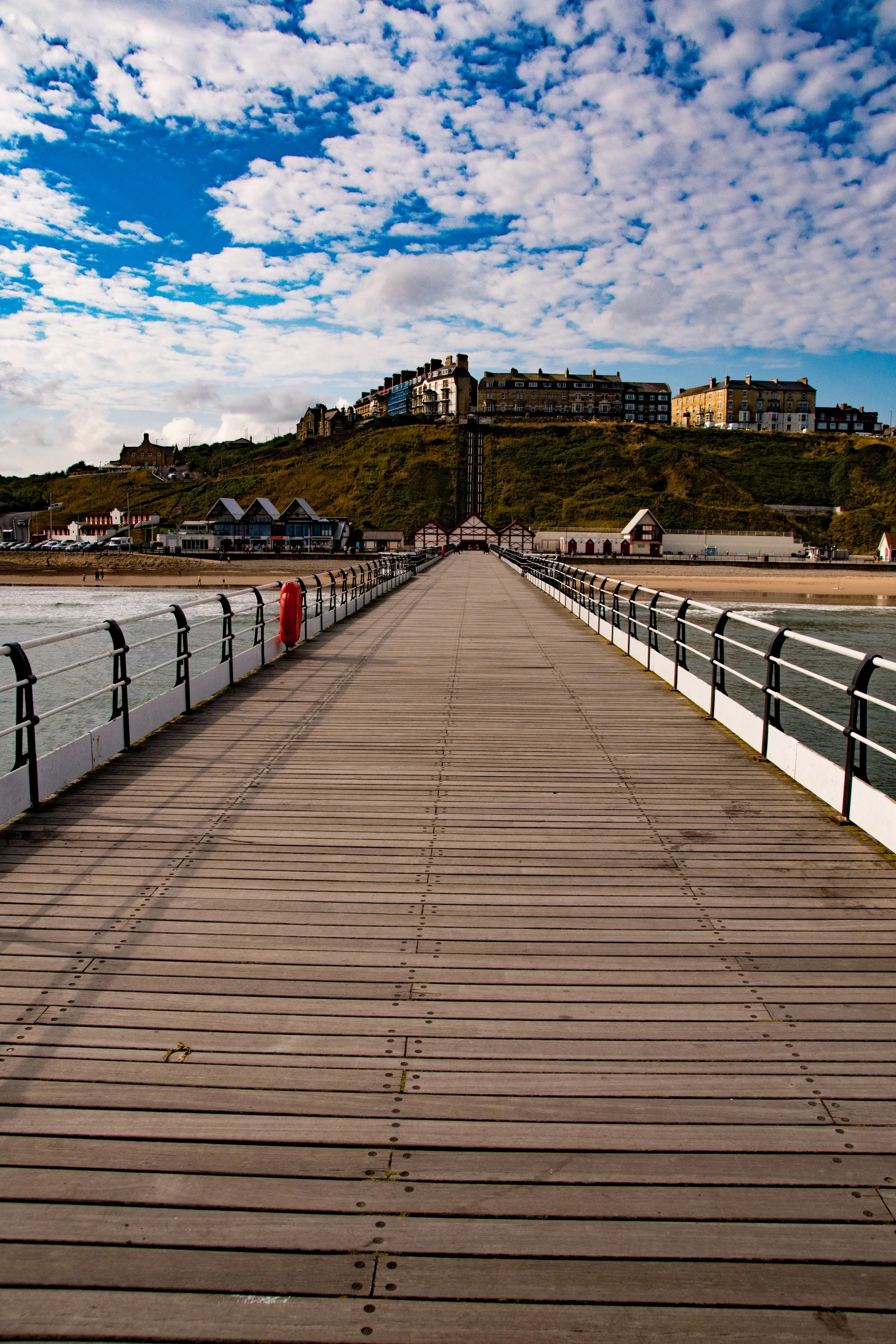 A wooden pier extending over water towards a hill with houses on top, under a partly cloudy sky.