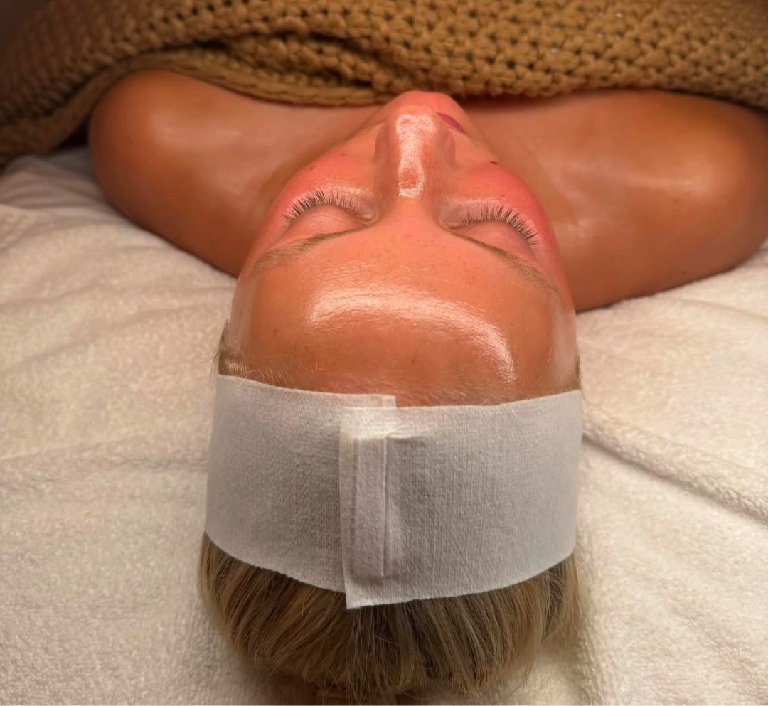 Person receiving a facial treatment with a headband, lying down with closed eyes and covered with a brown blanket.