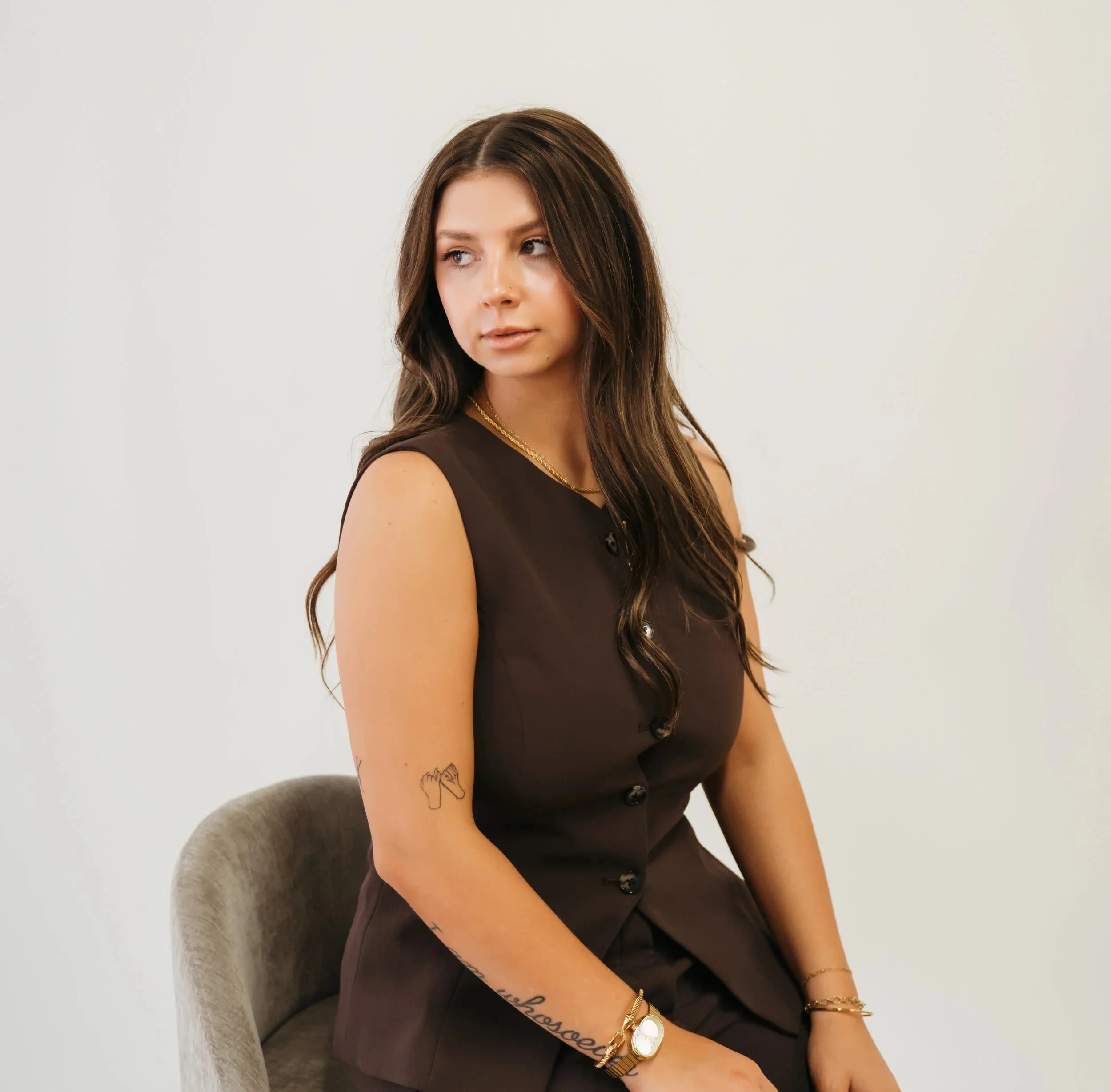 A woman with long wavy brown hair, wearing a sleeveless dark brown dress, sitting on a beige chair against a plain white wall, looking off to the side.