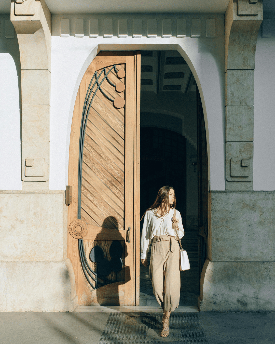 A woman with long hair wearing a white blouse and beige pants walks out of a large wooden doorway, casting a shadow on the door. The doorway has decorative metalwork and is part of a building with stone accents.