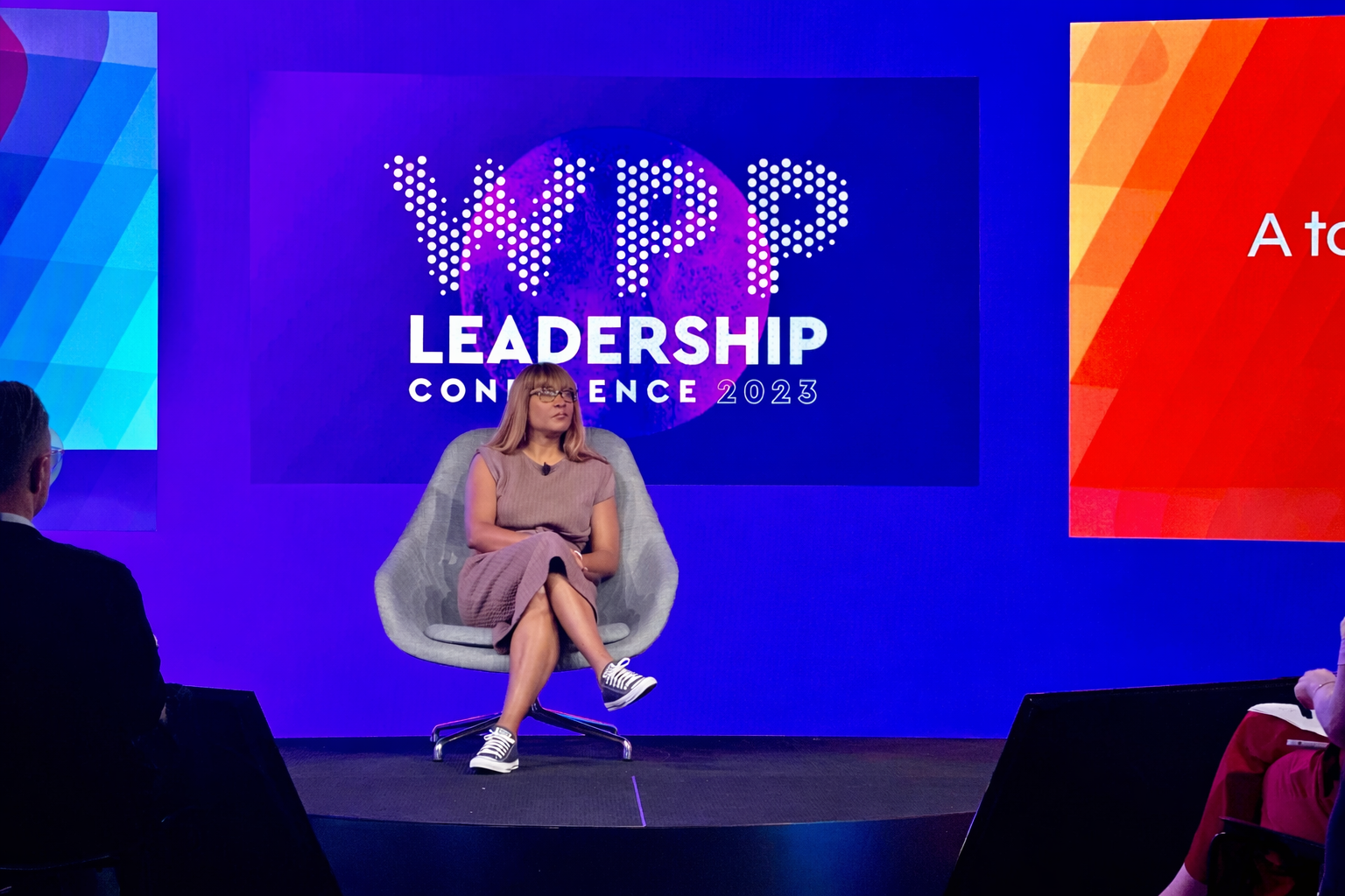 Woman in a brown dress and glasses sits centered on a stage chair at the WPP Leadership Conference 2023, framed by a large purple and blue WPP logo screen behind her, with the audience visible in the foreground.