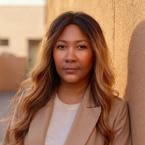 Portrait of a woman in a light tan blazer standing against a sunlit urban wall, looking directly at the camera with a calm, confident expression.