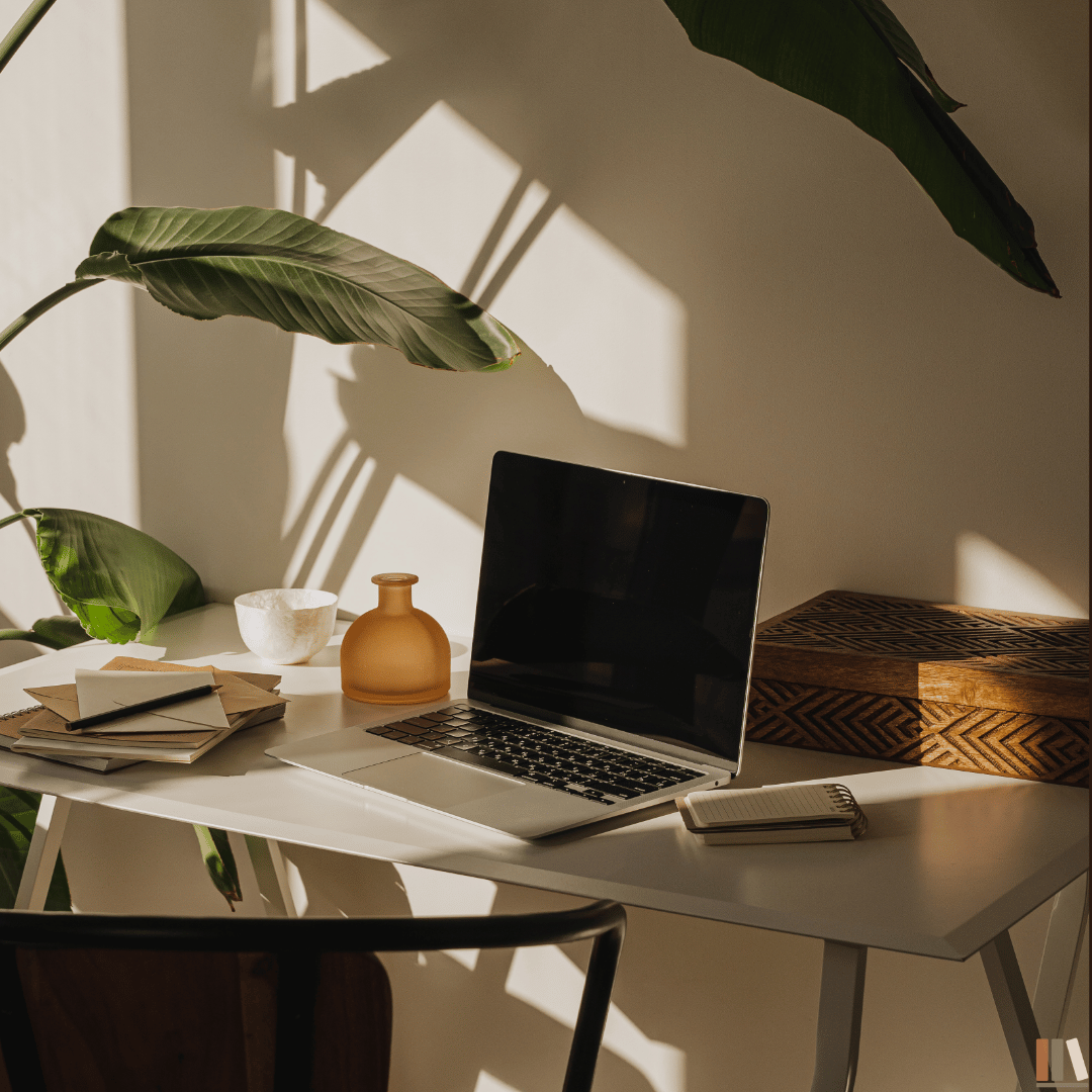 A white desk with a laptop, notebooks, a pen, a small ceramic bowl, a brown vase, and a rectangular decorative box, with large green leaves casting shadows on the wall behind.