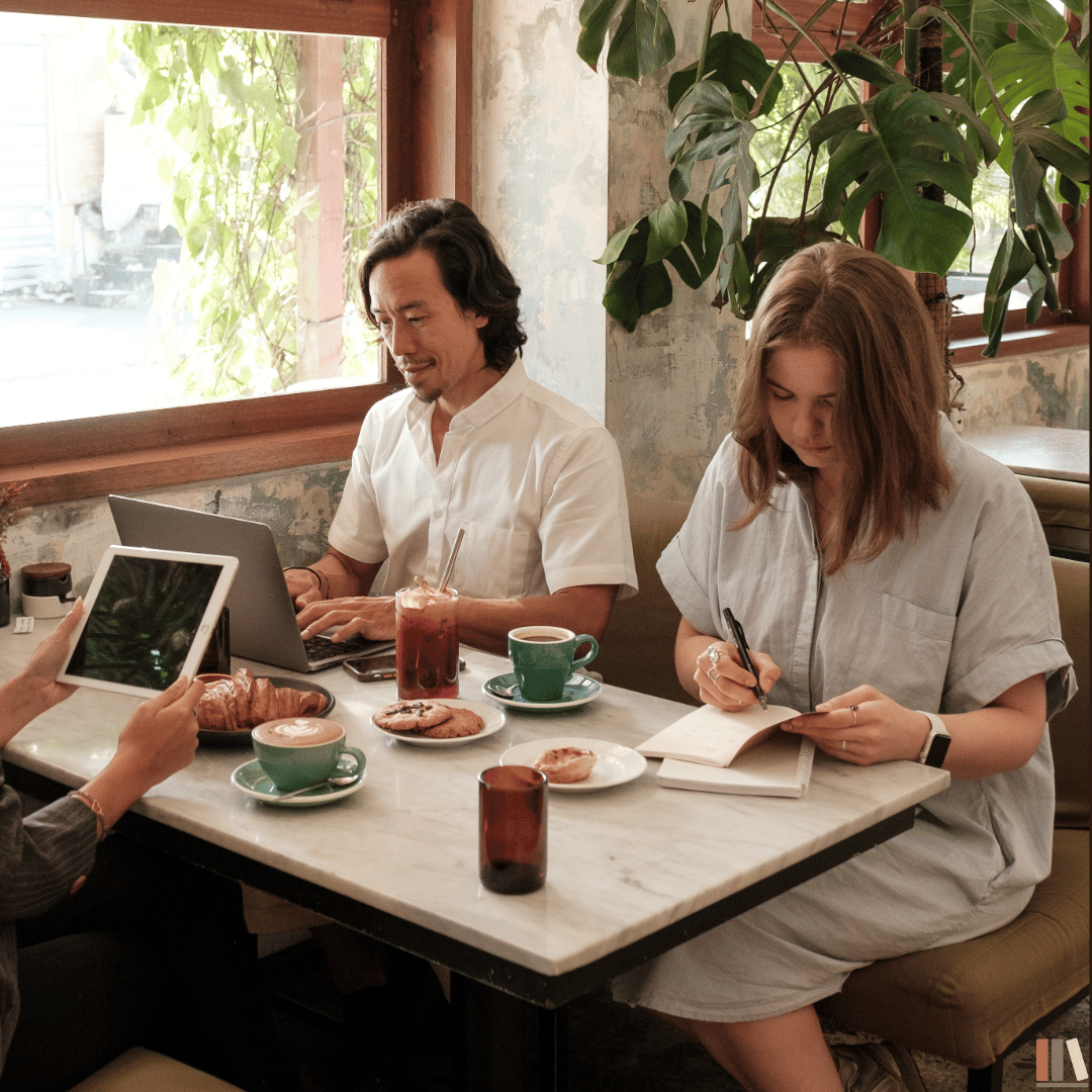 Two women and one man sitting at a table in a cozy café with large windows, writing and working on their devices surrounded by coffee cups, plates with snacks, and a potted plant.