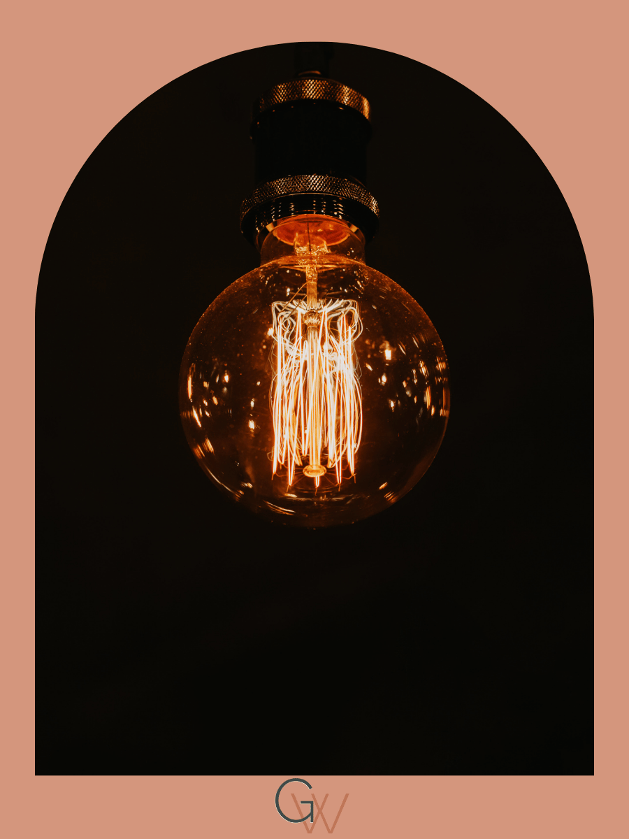 Close-up of an illuminated vintage Edison light bulb with filament visible, hanging against a dark background, framed with a peach-colored border.