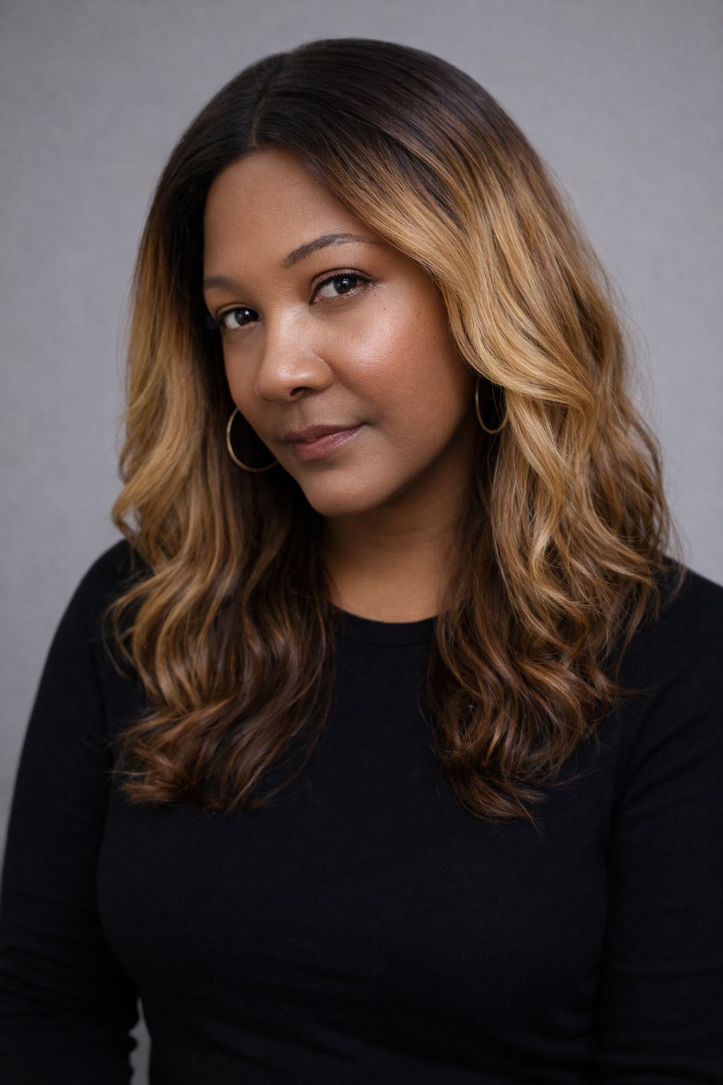 A woman with medium skin tone and wavy, shoulder-length hair with highlights, wearing a black top and hoop earrings, poses against a gray background.