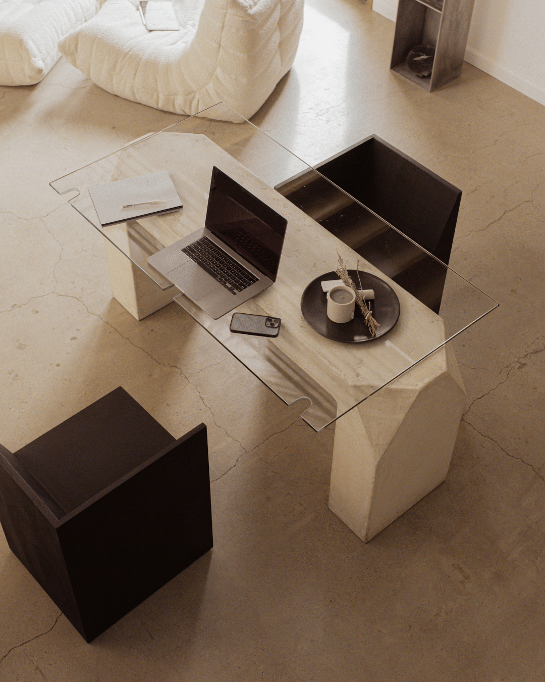 A modern dining table with a glass top, black chairs, a silver laptop, a smartphone, a black tray with a white mug and dried flowers, and a closed notebook with a pen inside. The table is in a minimalistic living space with beige floors and neutral furniture.