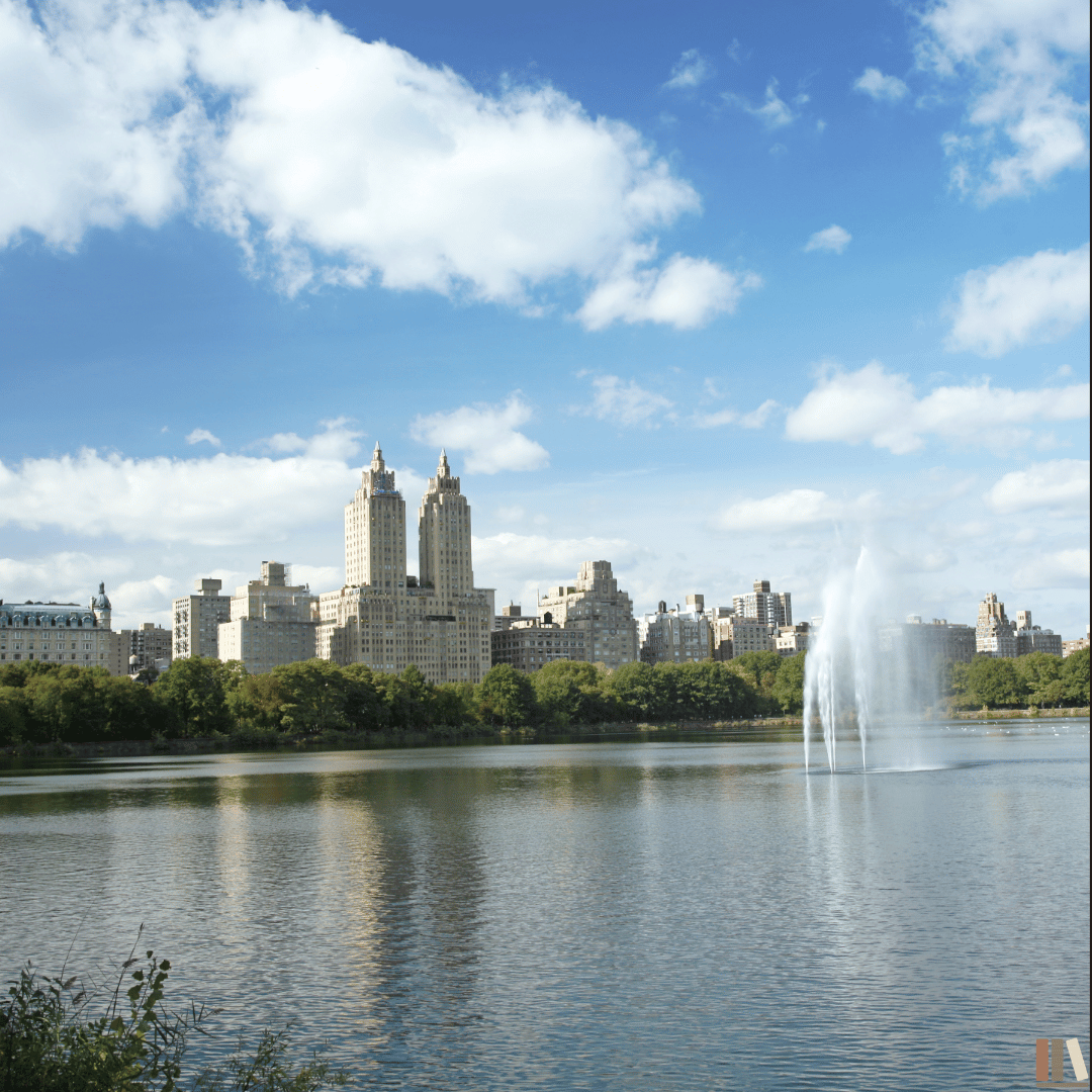 City skyline with tall buildings and a fountain in a lake, green trees, and a partly cloudy sky.