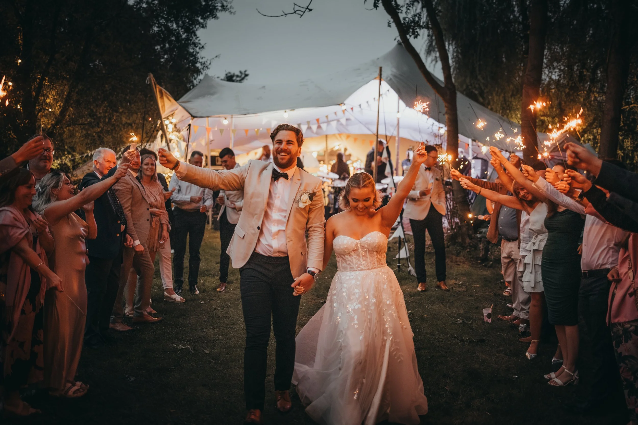 A newlywed couple walking hand in hand through a celebration surrounded by friends and family holding sparklers at night, under a large white tent decorated with string lights and bunting.
