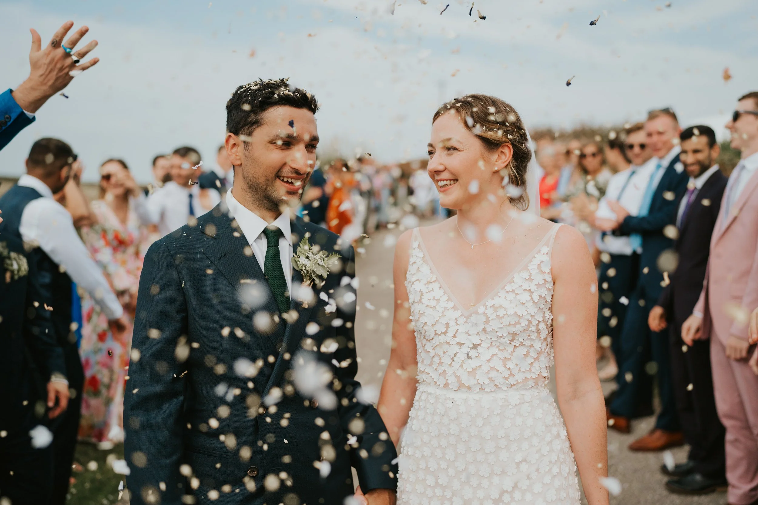 Bride and groom smiling amid confetti at Cornish outdoor wedding with guests looking on.
