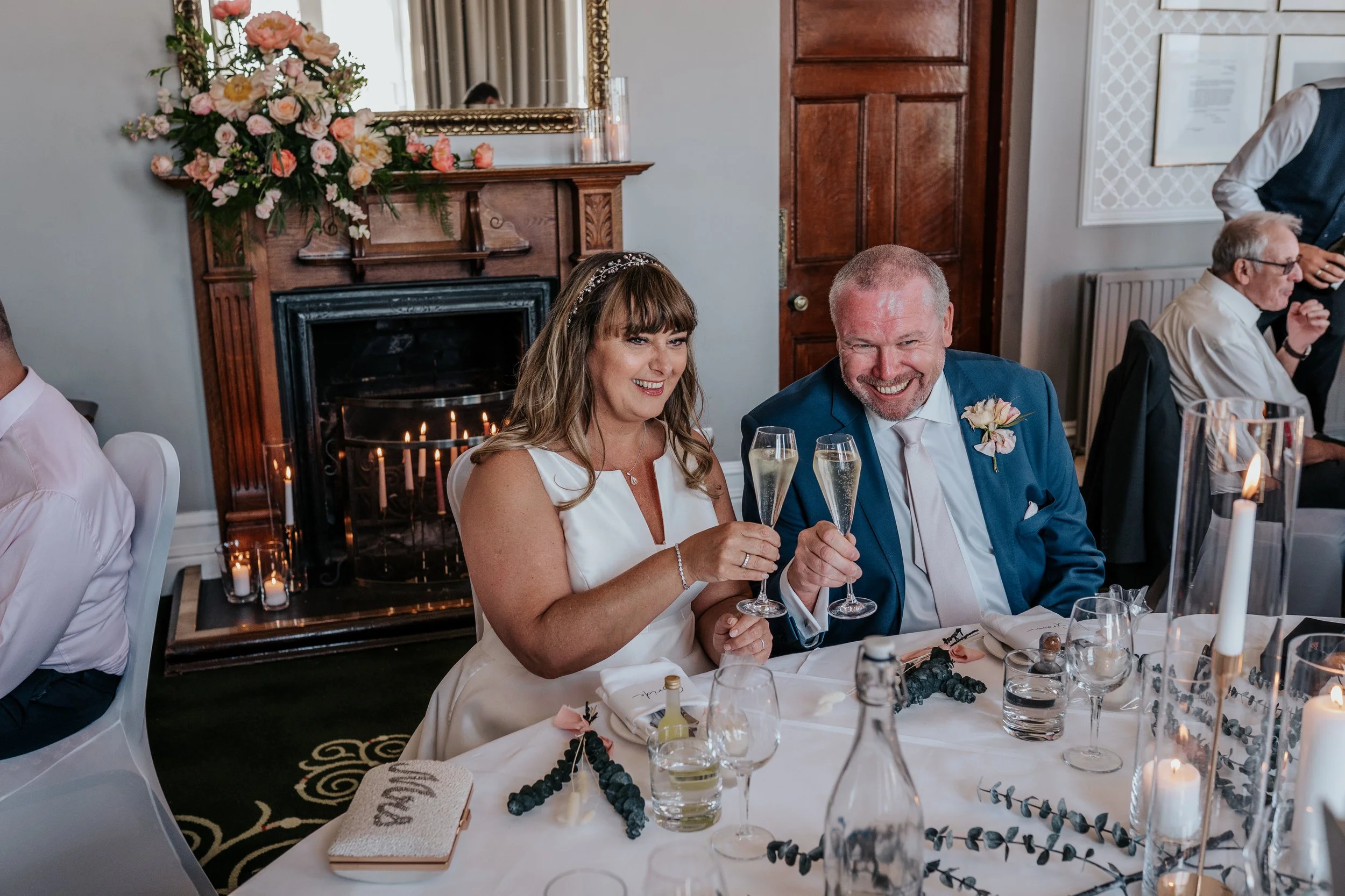 A bride and groom toasting with champagne glasses at a wedding reception table, decorated with candles and flowers. The room features a fireplace and a mirror above it.