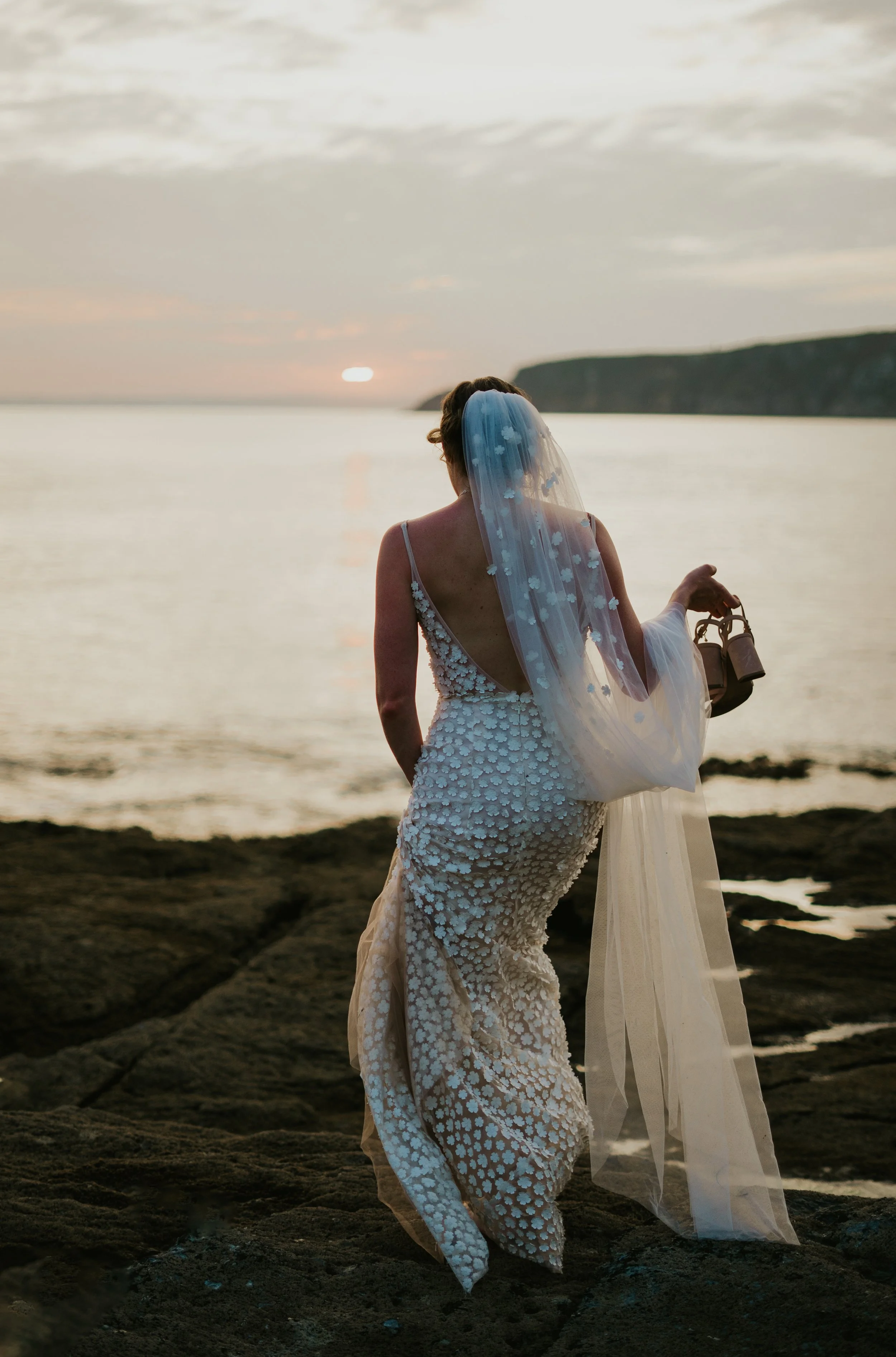 Bride in a wedding dress and veil standing on rocks by the ocean, in Cornwall, at sunset, holding shoes.