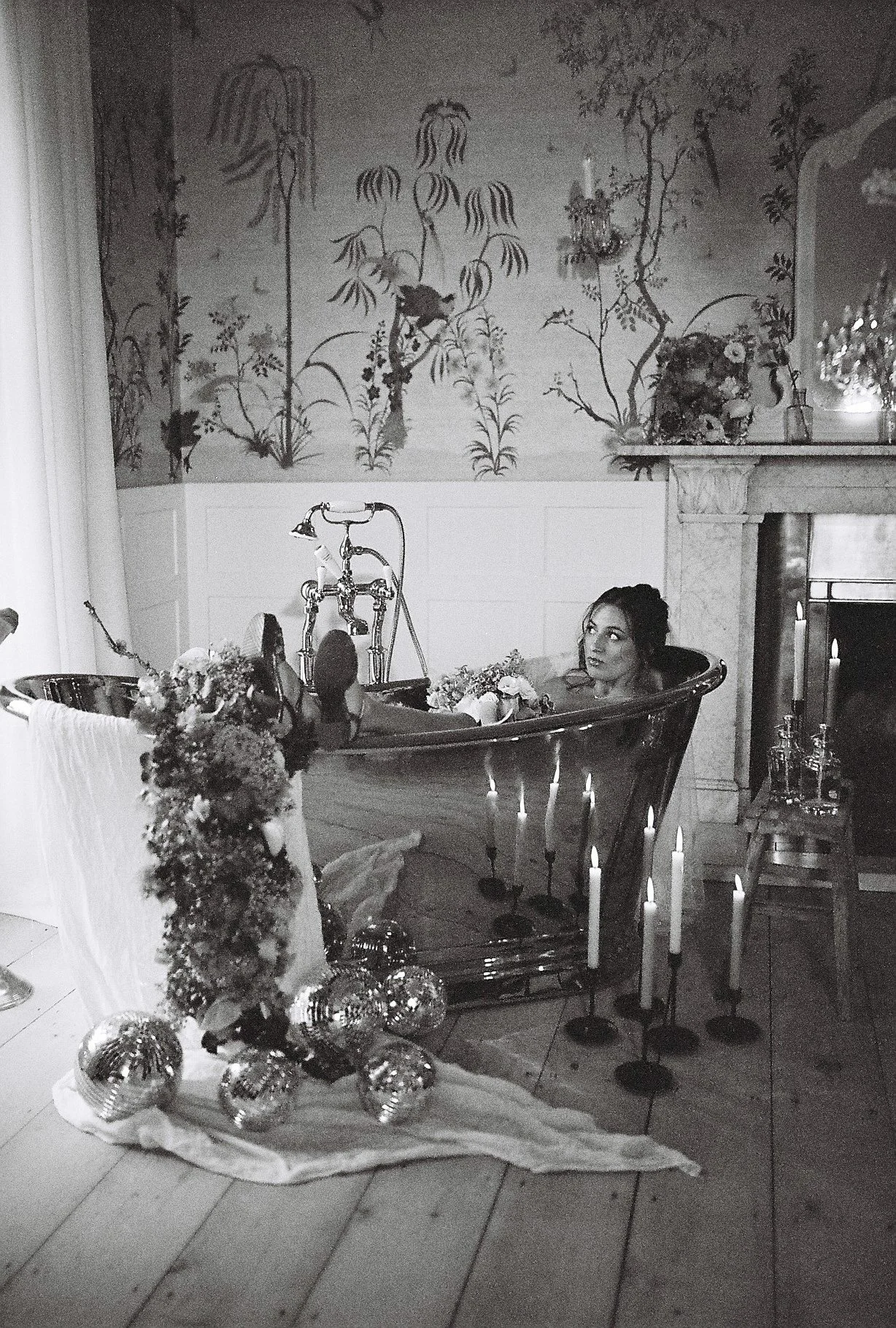 A woman is in a vintage bathtub filled with flowers, in a decorated room with candles and a floral wallpaper background.