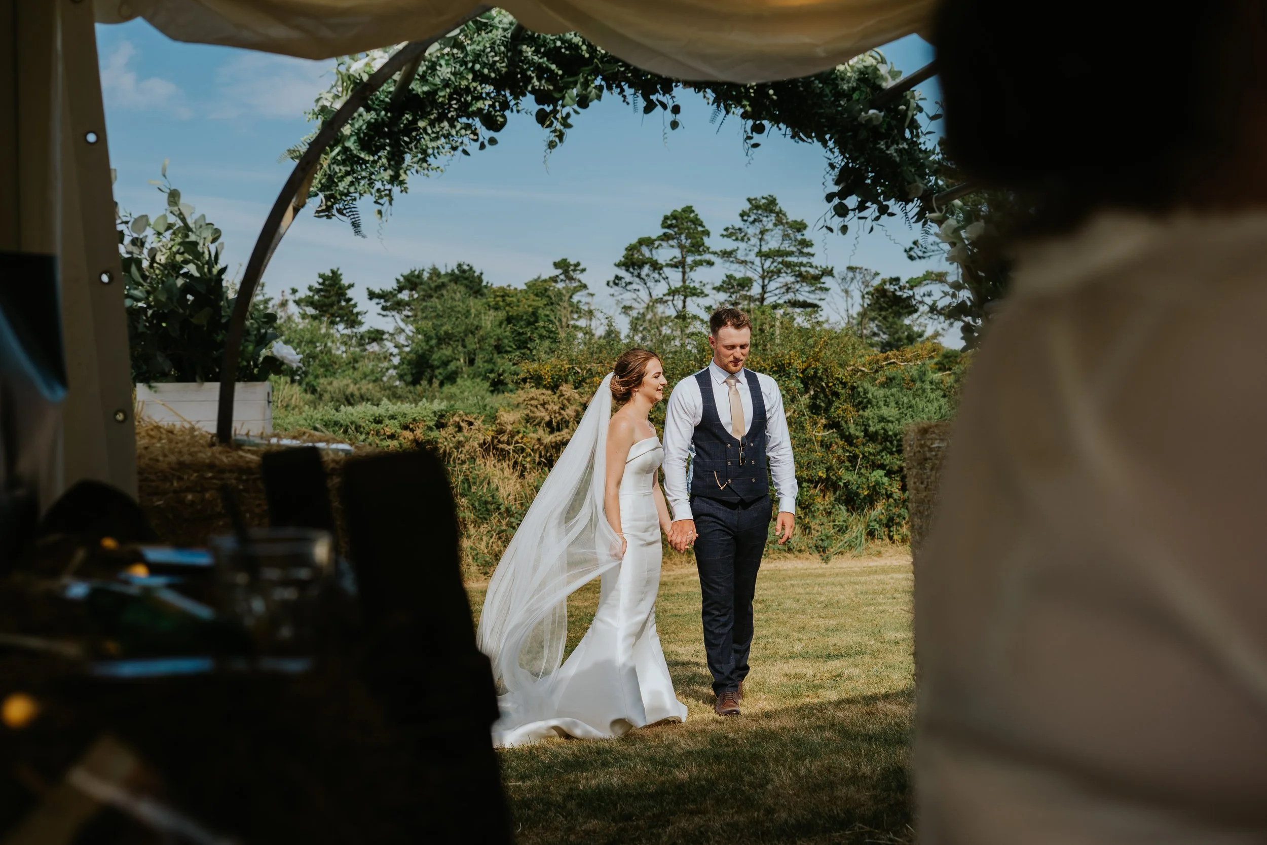 A bride and groom holding hands and walking outdoors on their wedding day, framed by a tent or canopy with greenery, trees, and a partly cloudy sky in the background.
