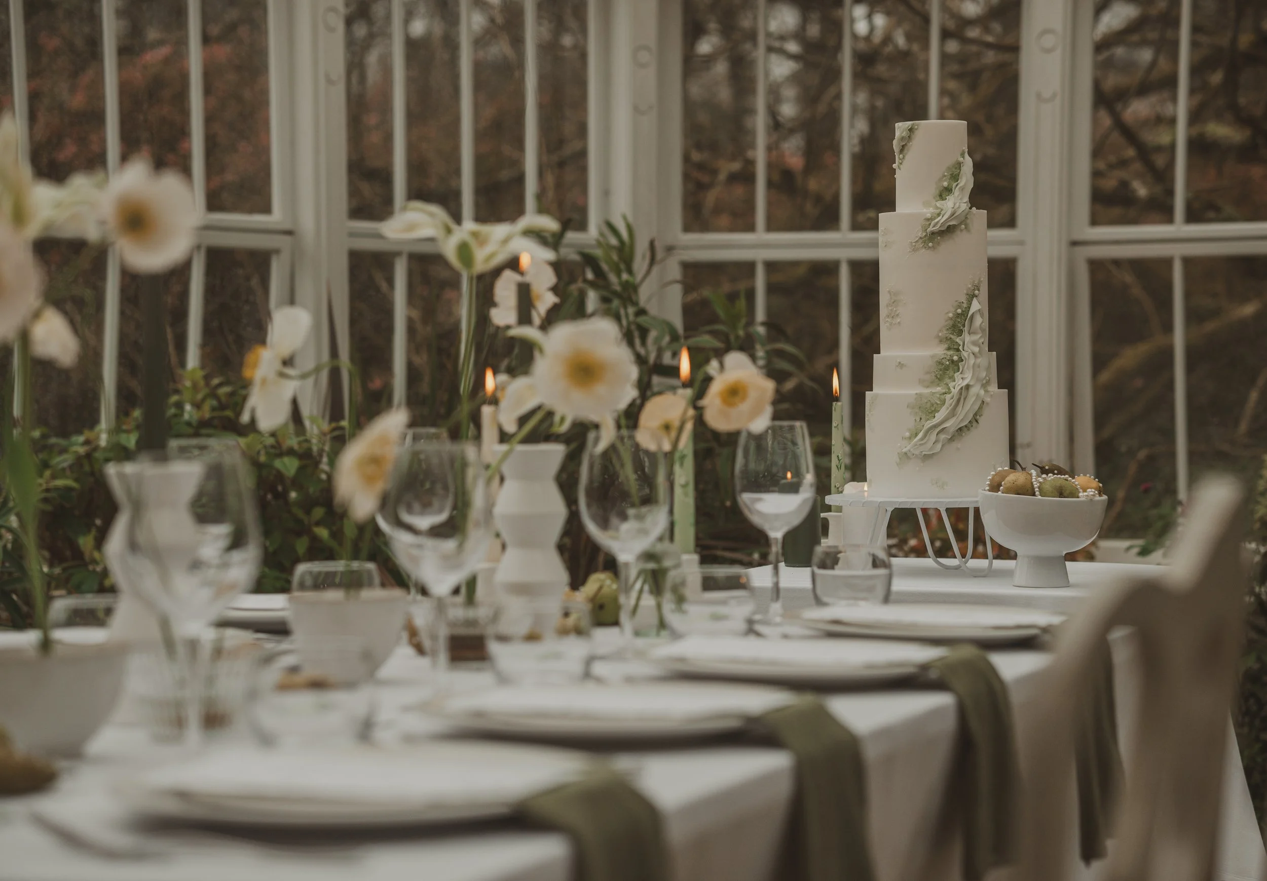 A decorated wedding reception table set with wine glasses, floral centerpieces, a white tiered wedding cake with green accents, and bowls of desserts, inside a bright conservatory with large windows and a view of autumn trees outside.