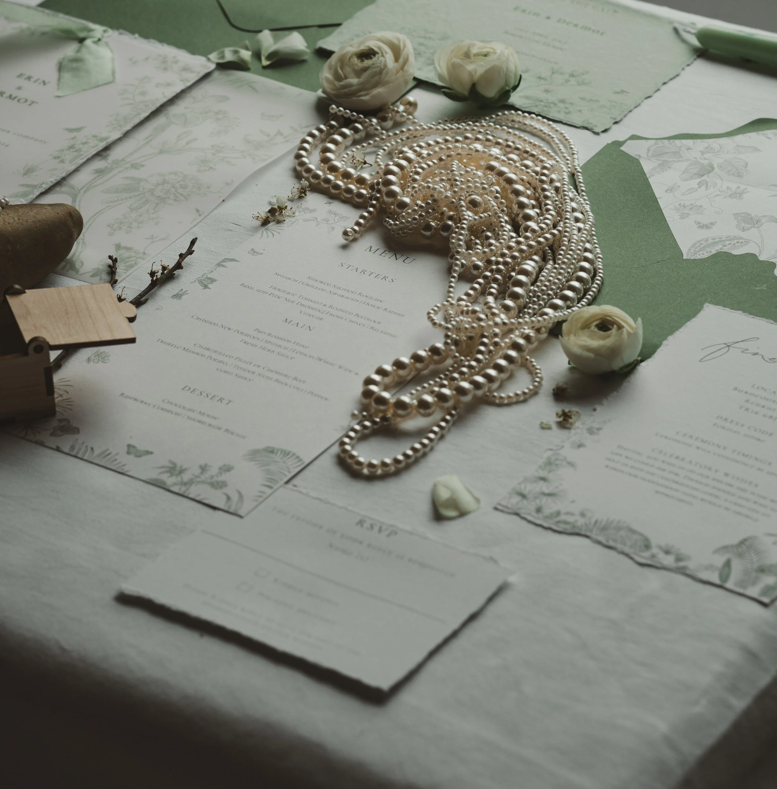 Wedding invitation suite with a menu, pearl necklaces, white roses, and greenery on a light-colored table.