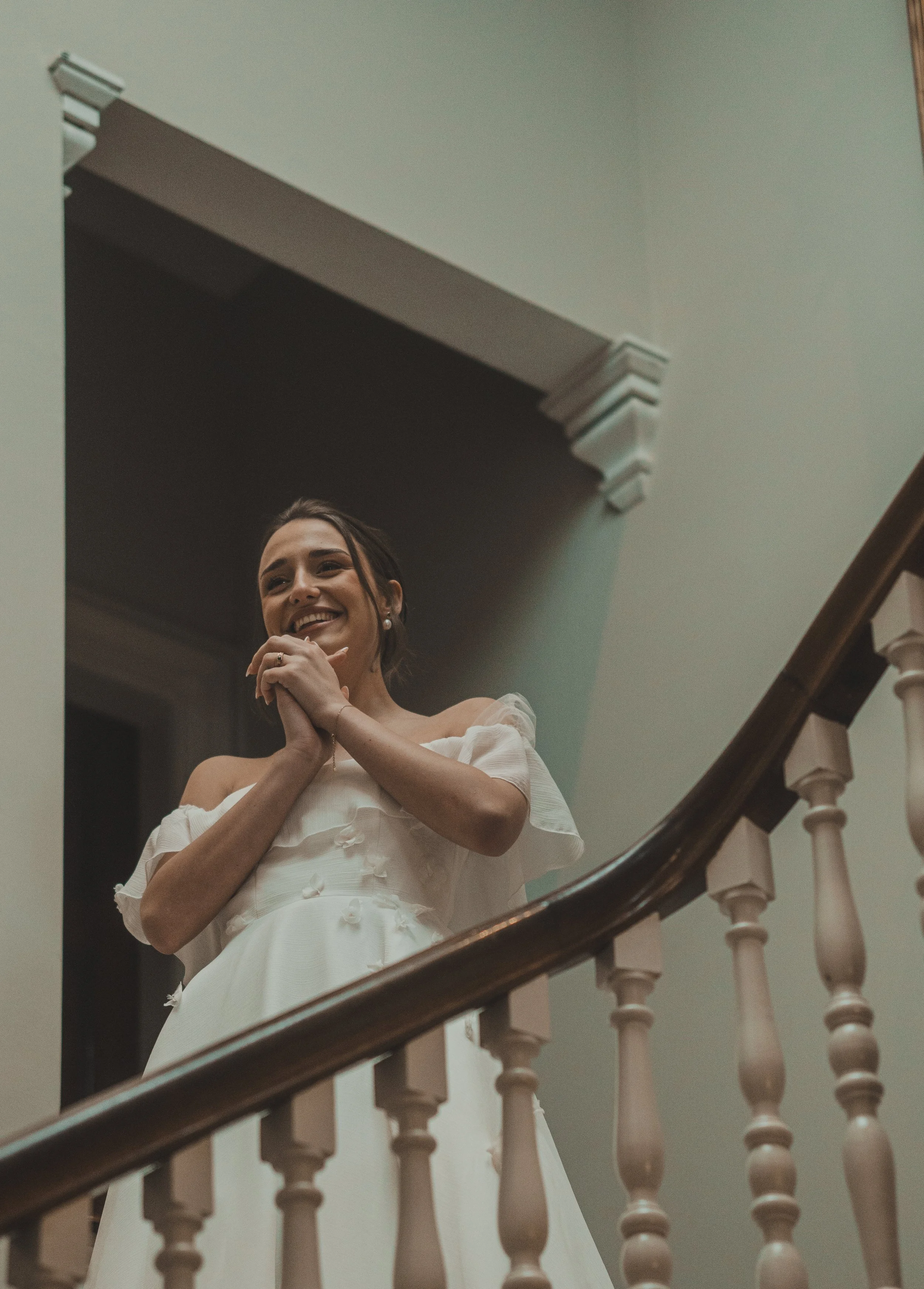 A woman in a white wedding dress standing on a staircase, smiling and holding her hands together near her face.
