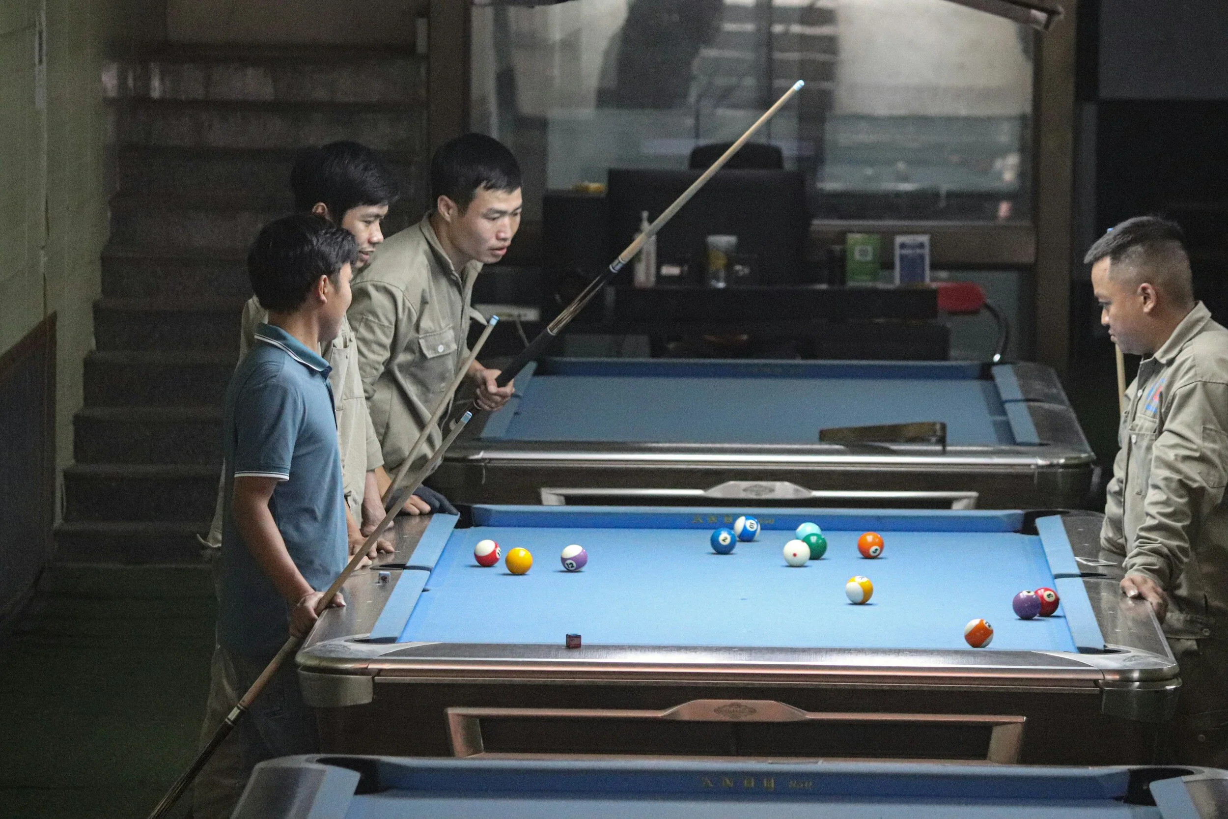 Four men playing pool at a billiards hall with multiple blue-felt tables and scattered billiard balls.