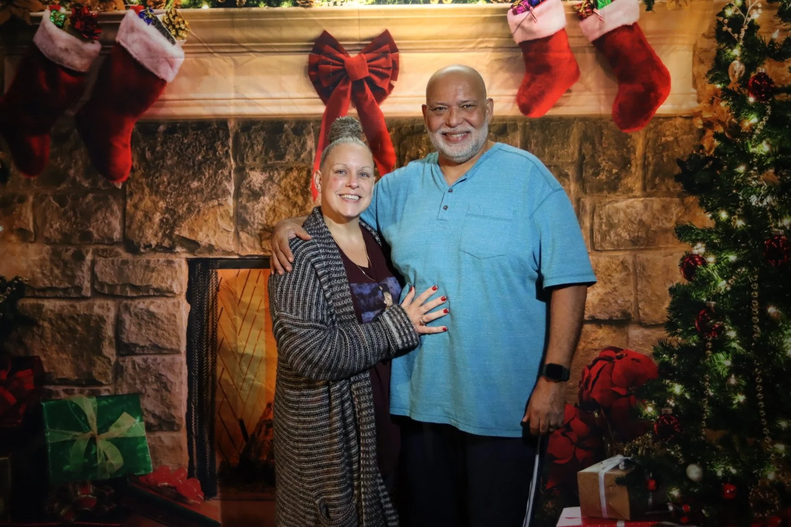 A smiling couple embracing in front of a Christmas fireplace scene, decorated with stockings, ornaments, and a Christmas tree.