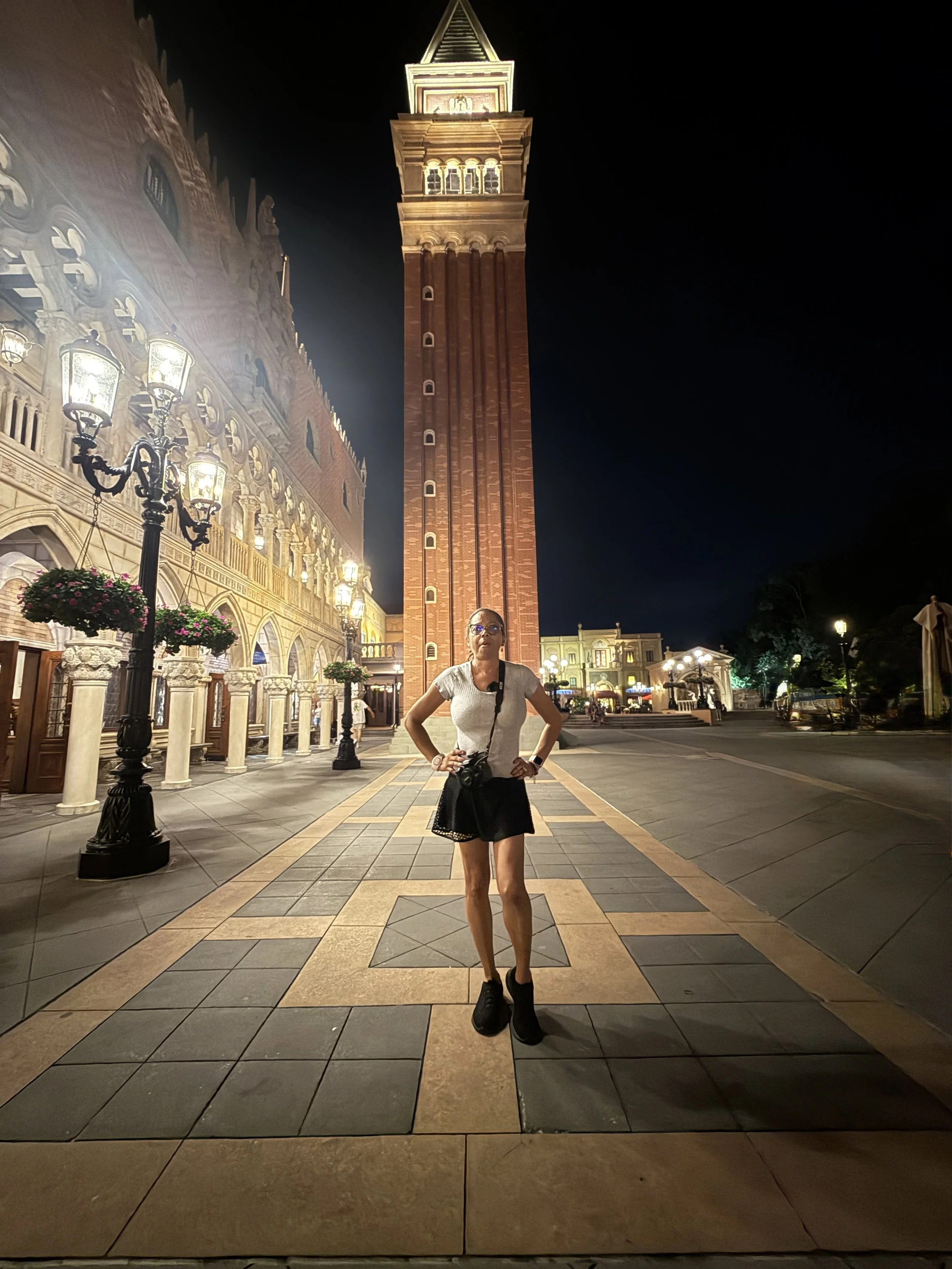 A woman standing with hands on hips on a tiled street at night, with a tall clock tower and illuminated buildings in the background.