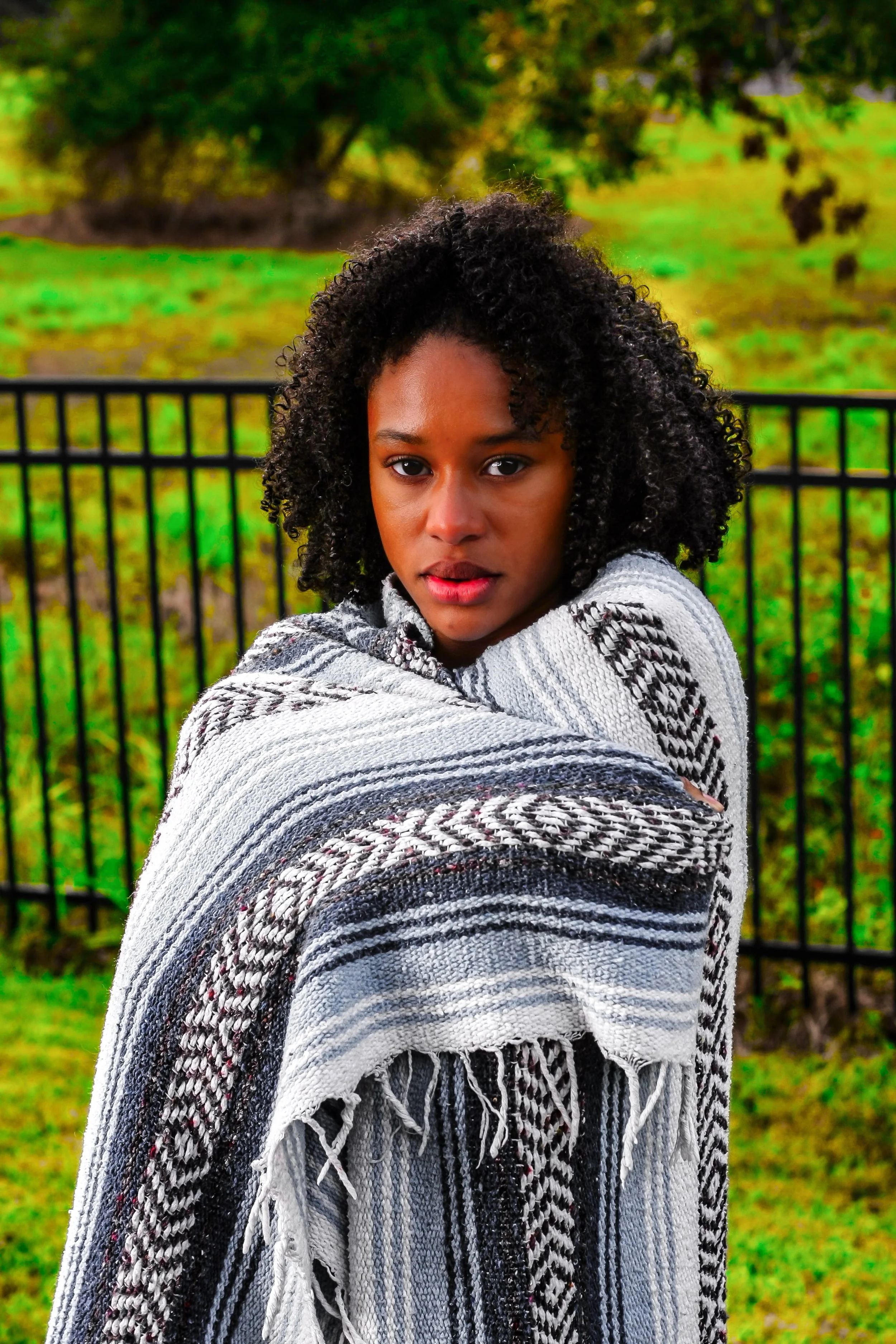 A young woman with curly black hair standing outdoors wrapped in a black and white patterned blanket, with green grass and trees in the background.